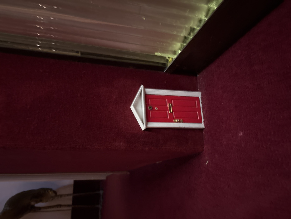 The image displays an indoor scene, likely a hotel hallway or room, characterized by dark red carpet covering both the floor and the lower portion of a wall or column. Centrally positioned at the base of this wall is a miniature, bright red door with white trim. The door features a small white triangular pediment above its frame. Essential details on the red door include a gold door knocker, a horizontal gold letterbox slot, and a gold doorknob. To the left, a darker area of the carpeted floor recedes into the background, where the lower legs and feet of a slender, dark, possibly metallic, statue or decorative object are partially visible. To the right of the miniature door, a large window or glass panel is present, partially obscured by light-colored, vertically hanging translucent curtains. The bottom frame of the window is dark. The scene is dimly lit, with ambient light filtering through the curtains on the right. No people or overt actions are depicted. The visual evidence within the image does not contain specific identifiers to determine a precise location within Paris, France.