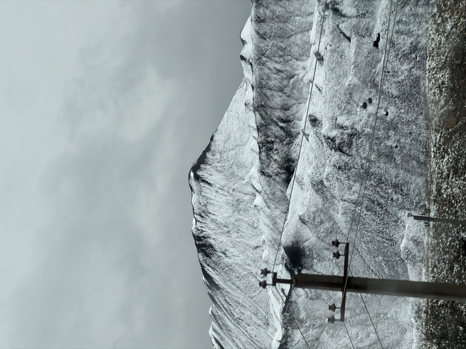 The image displays a mountainous winter landscape in the vicinity of Terjola, Georgia, under an overcast sky. In the upper background, a prominent, snow-covered mountain peak with exposed dark rock formations dominates the horizon. Below this peak, a series of undulating hills and lower slopes are heavily dusted with snow, revealing irregular patches of darker, exposed terrain or sparse vegetation.
In the foreground, positioned on the left, a vertical utility pole, dark in color and appearing to be constructed from concrete, features three horizontal crossarms. Each crossarm supports two brown or reddish-brown insulators, from which multiple parallel power lines extend horizontally across the frame, partially obscuring the midground hills. A smaller, similar utility pole is visible further back and to the right. The immediate bottom edge of the frame shows a strip of dark ground or low-lying vegetation also covered with a light layer of snow. The sky is uniformly light grey and cloudy. No individuals, animals, or specific actions are present in the scene.