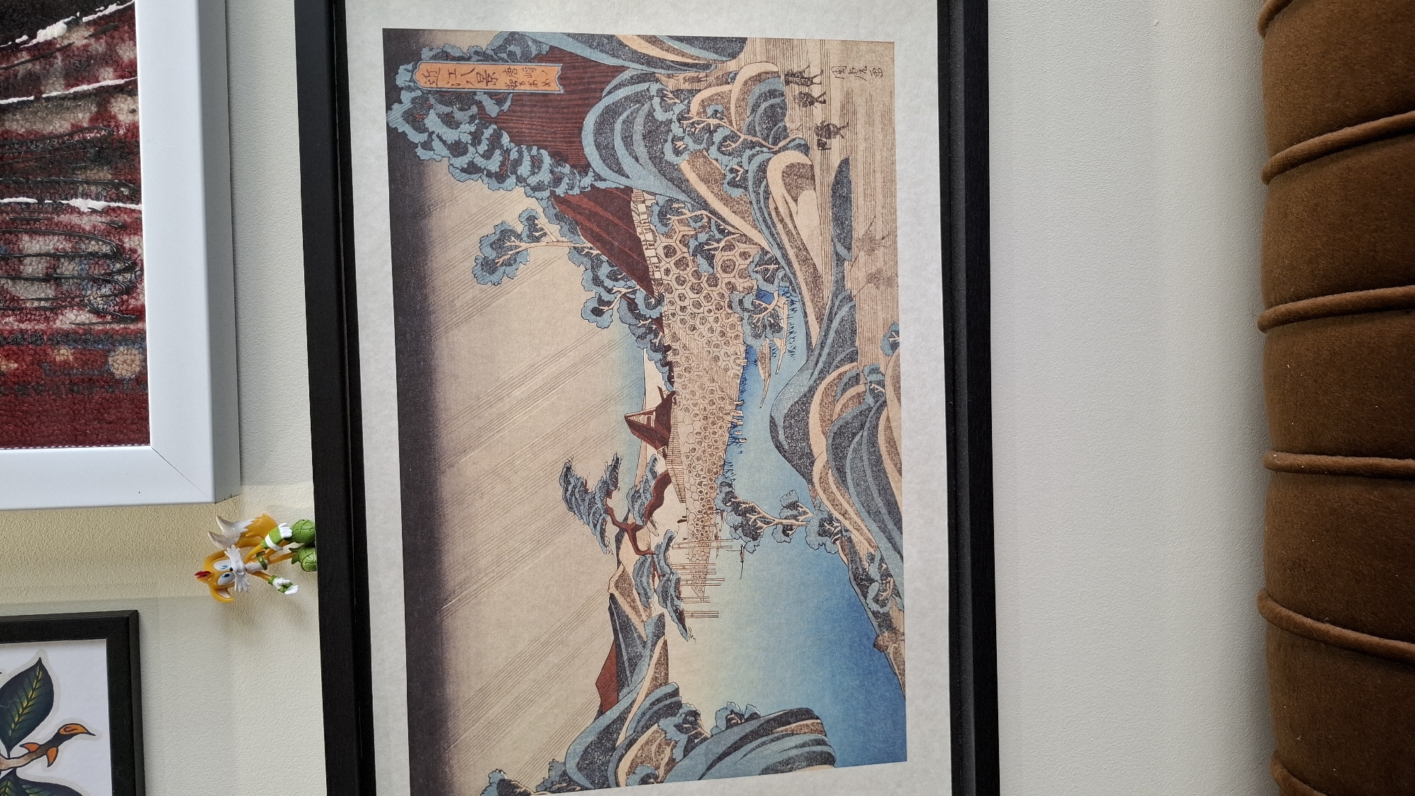 The image shows a framed Japanese woodblock print hanging on a white wall. The print depicts a landscape scene with rolling hills, a body of water, trees, and small figures of people walking. The print has a reddish-brown frame with a black mat. Above and to the left of the woodblock print is another framed artwork, partially visible, and a small figurine of a character resembling Tails from Sonic the Hedgehog. The bottom of the image shows the top of a brown upholstered furniture item. The scene is static and does not depict any specific location in Plovdiv, Bulgaria.