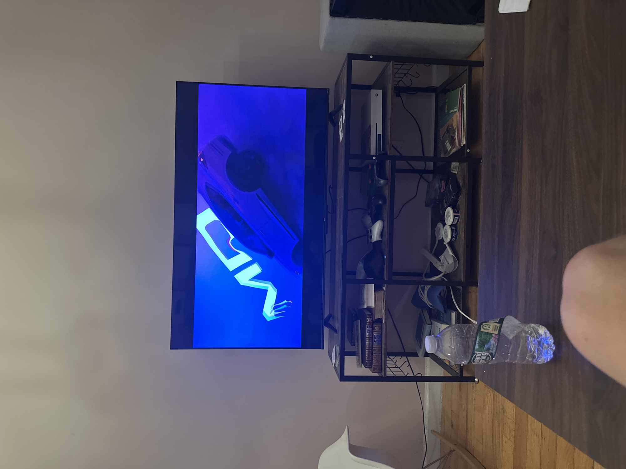 A television is mounted on a wall beside a black metal shelving unit containing an Xbox, video game controllers, and other items. A plastic water bottle is on the table in front of the shelf. A white chair is visible in the bottom left corner of the frame. The location is likely a residential interior in New York, United States of America.