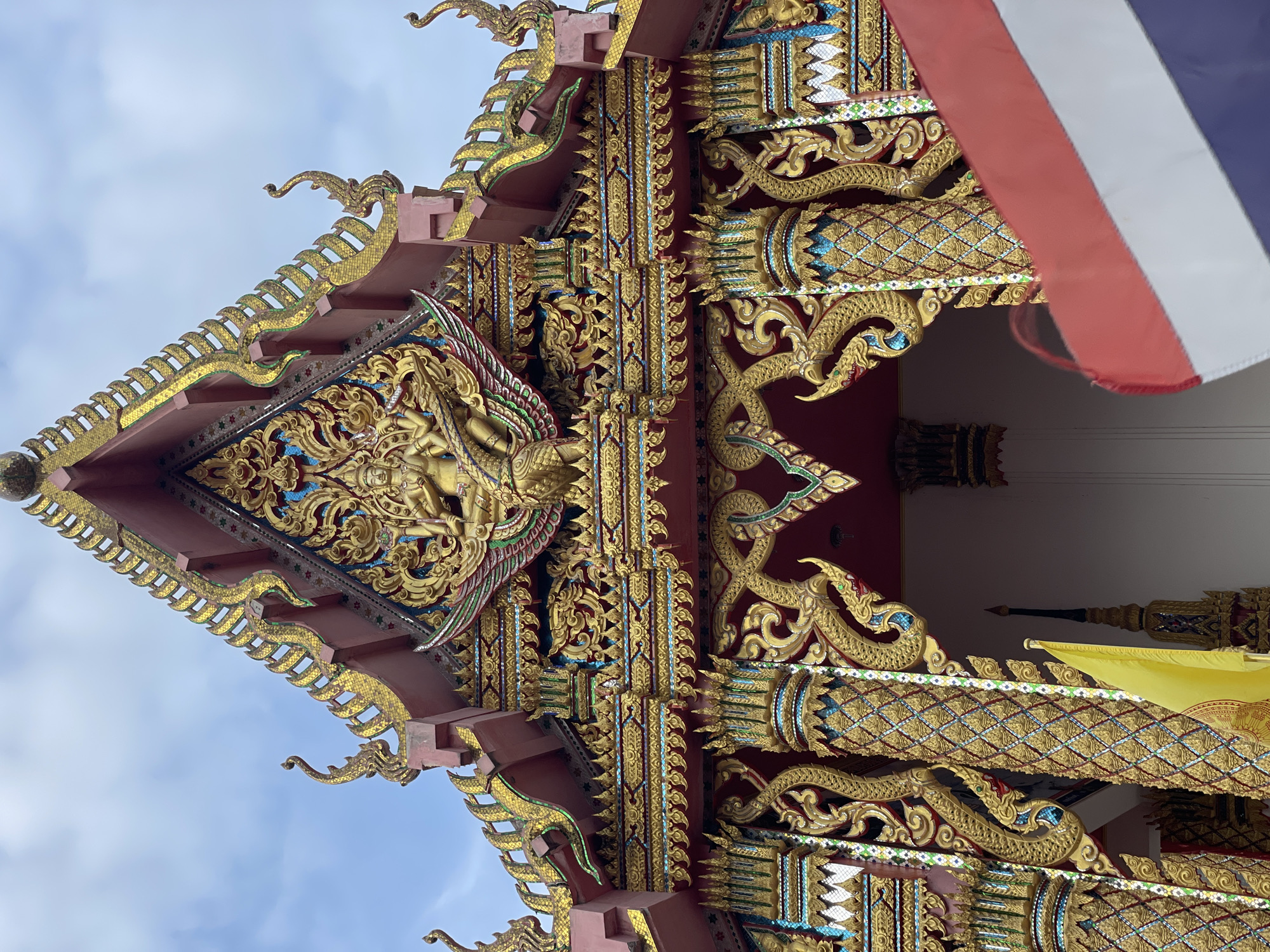 A close-up, low-angle shot captures the ornate pediment of a temple in Pattaya City, Thailand. The architecture is adorned with intricate golden carvings, featuring mythical creatures and celestial beings. Dominating the upper center is a sculpture of a multi-armed deity riding a bird-like creature, surrounded by elaborate floral motifs and serpentine designs. Flanking the central sculpture are decorative golden finials that follow the steeply pitched roofline. The temple's facade is painted a deep red, contrasting with the abundant gold embellishments and touches of blue and green tiles. In the foreground, partially obscuring the lower portion of the temple, are two flags: a bright yellow flag on the left and a blurred Thai flag on the right, displaying red, white, and blue stripes. The sky above is overcast with a pale blue hue. The overall impression is one of rich cultural and religious artistry characteristic of Thai temples.