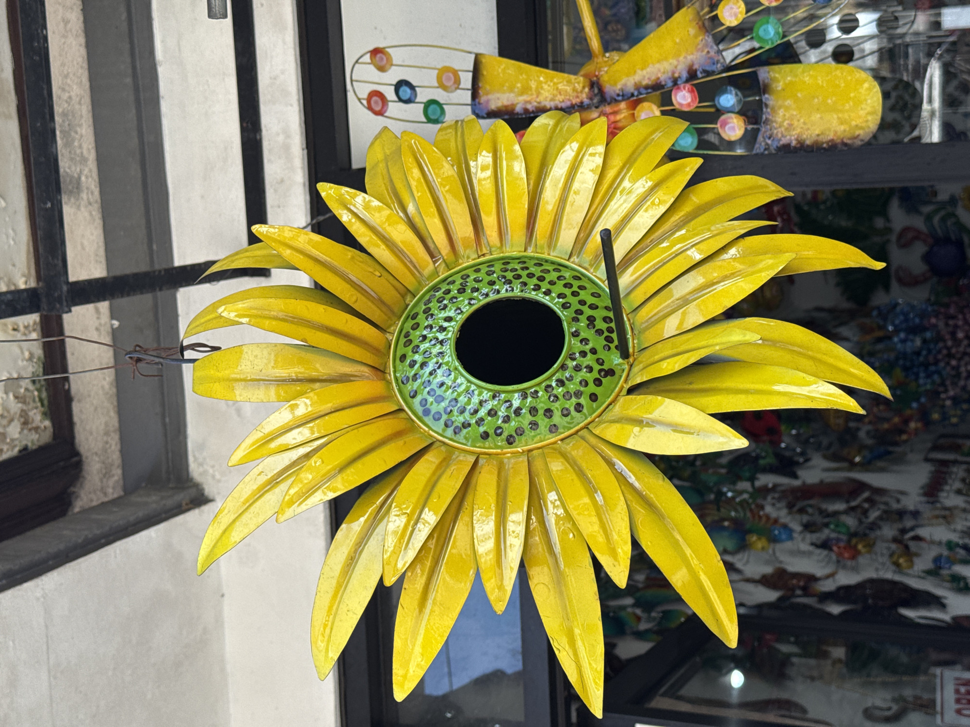 A close-up, eye-level, outdoor shot shows a bright yellow, metal sunflower-shaped birdhouse hanging from a wire. The sunflower has a green center dotted with dark brown spots and a black circular opening for birds. The petals are elongated and curved, with a shiny, reflective surface that shows droplets of water. To the right of the sunflower, a colorful metal butterfly decoration with multicolored glass orbs hangs. The background is blurred but appears to be a shop or market stall with various items displayed behind a window, including a red and white "OPEN" sign at the bottom left. The setting suggests a decorative item displayed for sale in a commercial area, possibly in Ubud, Indonesia.
