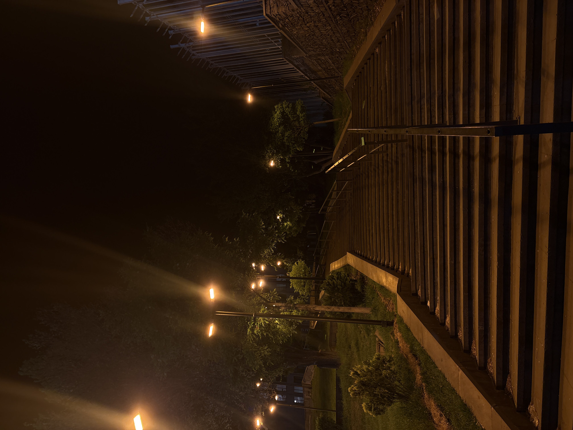 The image depicts an outdoor scene at night in Yerevan, Armenia. A set of concrete steps stretches upward to the right side of the image. The steps are illuminated by warm lighting, creating alternating bands of light and shadow. A metal railing is visible on the steps. To the left of the steps, a park area is visible, with trees and bushes sparsely lit by lampposts that cast an orange glow. The background is dark, suggesting a nighttime environment. A fence, composed of vertical concrete or stone pillars, is seen along the right, above the steps.