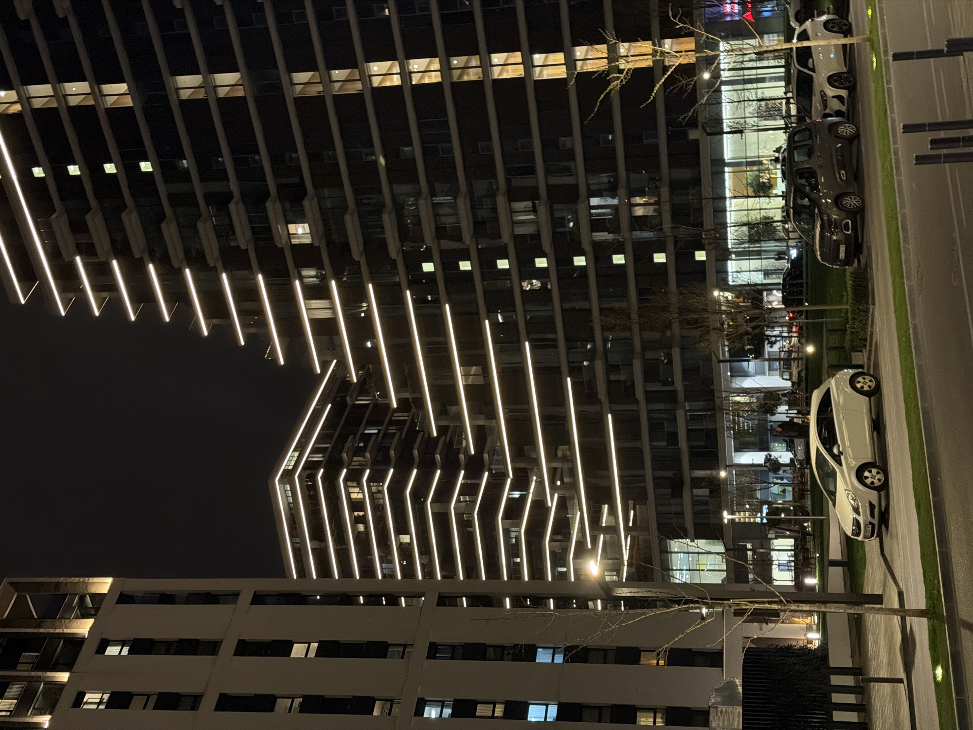 The image displays an urban night scene located within the Belgrade Waterfront complex in Belgrade, Serbia. The scene is dominated by three modern high-rise buildings under a dark, clear sky. On the left, a tall, light-colored residential building features vertical window arrangements, with some interior lights visible. To the right and extending into the background are two darker-toned, multi-story buildings, characterized by prominent horizontal bright white LED light strips that illuminate the edge of each floor or balcony. Numerous windows on these buildings show interior lighting, varying in color temperature from warm yellow to cool white.
At ground level, a paved street or roadway is present in the foreground, bordered by a grassy median strip and a sidewalk. Several bare-branched deciduous trees are sparsely distributed in the green areas and along the sidewalk, alongside some low shrubbery. Multiple modern streetlights provide white artificial illumination to the ground surfaces.
Three distinct vehicles are parked on the right side of the frame. A white two-door hatchback (Opel Corsa D) is parked diagonally on a paved section adjacent to a grassy area. Behind it, a dark-colored compact van (Citroën Berlingo) and a white SUV-type vehicle are parked parallel to each other. Faint human figures are discernible through the large, illuminated glass windows of the lower-level structures, indicating interior activity.