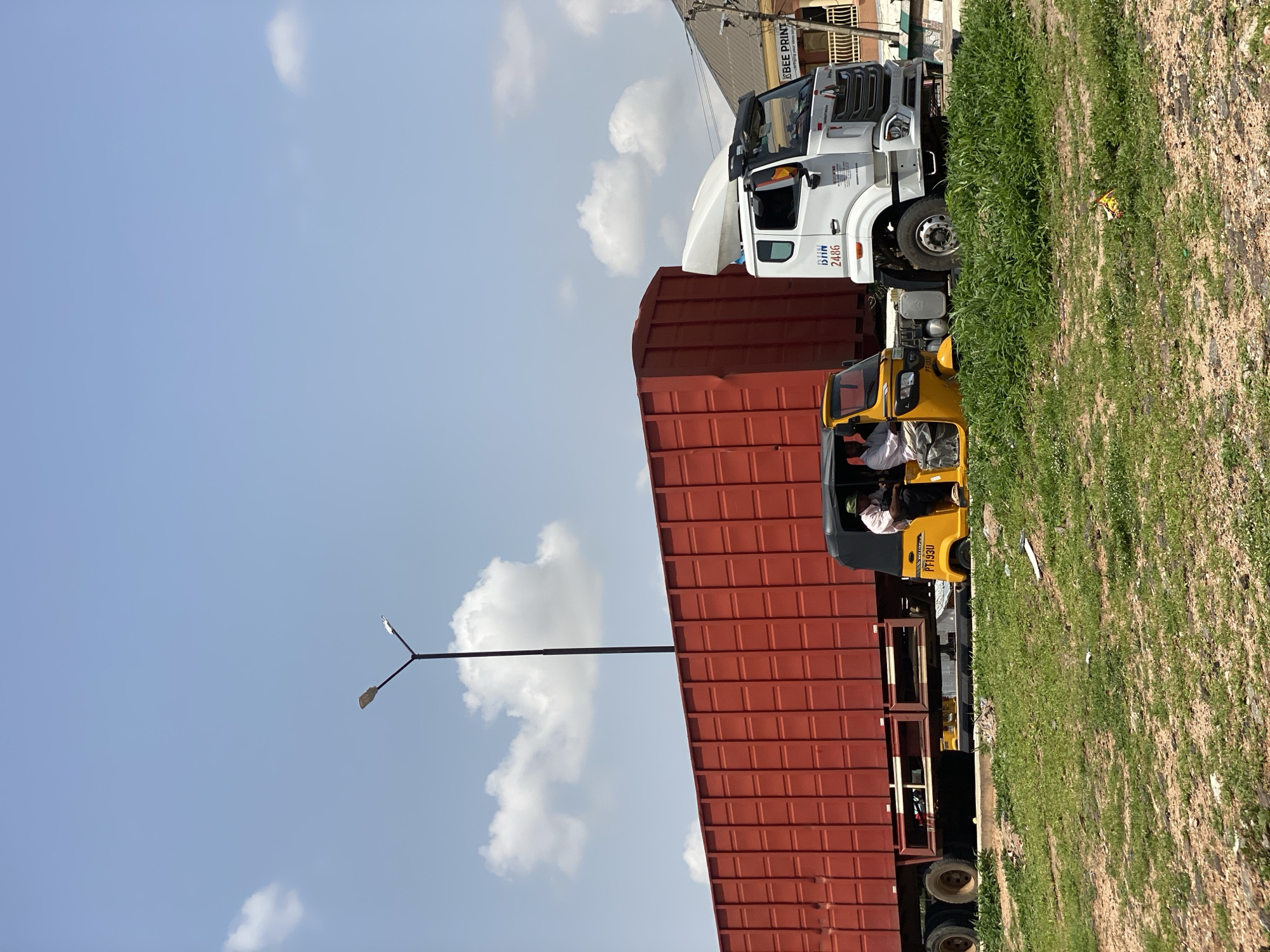 This image shows a street scene in Jalingo, Nigeria, under a clear blue sky with scattered white clouds, suggesting a bright, sunny day. The perspective is from a low angle, looking upwards.
The primary subjects are two vehicles: a large white truck and a yellow auto-rickshaw. The white truck is parked in front of a red-roofed structure, possibly a building or a large container. The truck has a portion of its hood open, revealing mechanical components. Visible text on the truck includes "BEE PRINT" on a sign above, and "DITH 2486" on the side of the cab.
In front of the truck and partially obscured by its trailer is a yellow auto-rickshaw. A person, likely the driver, is visible inside the rickshaw, wearing a light-colored shirt. The auto-rickshaw has a license plate that appears to read "PT 193U".
To the right of the vehicles is a patch of tall green grass, growing on uneven, dry ground, with some scattered debris. A black lamppost with two lights is visible in the sky, positioned above the red roof. The overall impression is one of a commercial or transportation hub, possibly a marketplace or a loading area, during daylight hours.