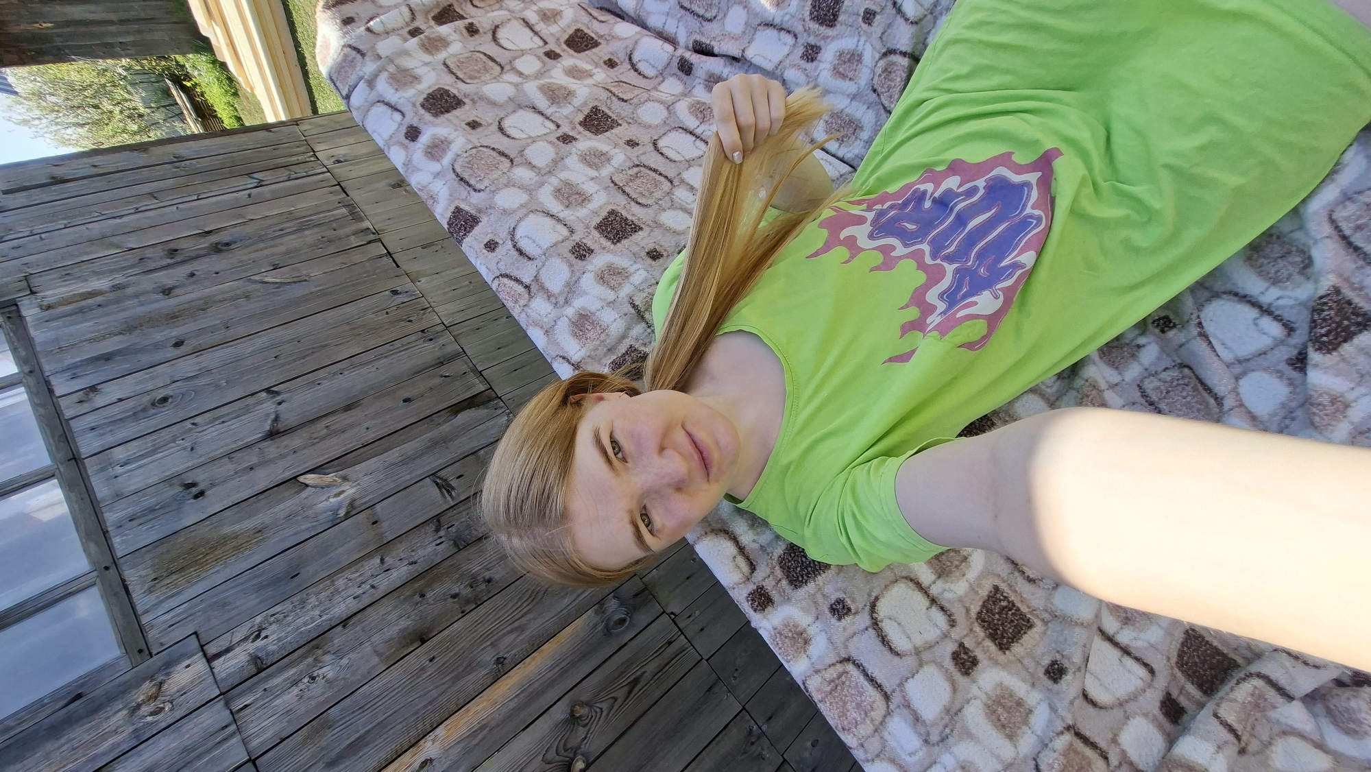 A person lies on a patterned fabric on a wooden deck, in what could be Большой Букор, Russia, based on visual clues. The person has blonde hair and is wearing a neon green shirt with a purple design. The deck is made of weathered wooden planks and has a window. Outside, there's foliage and sky visible.