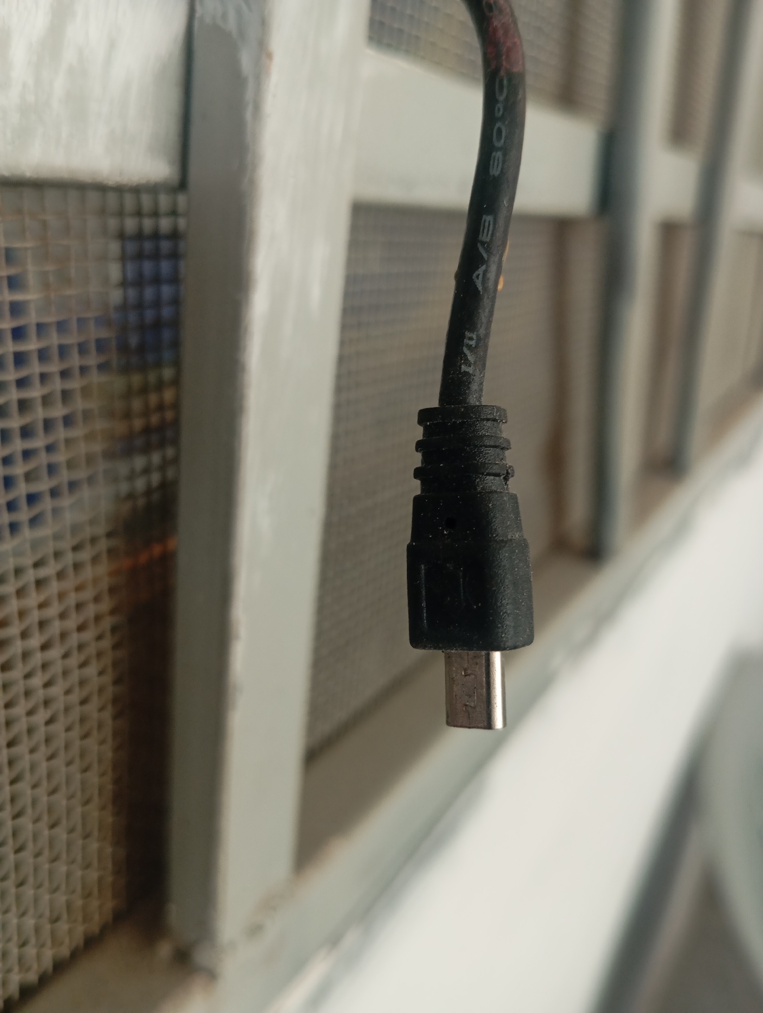 An outdoor, close-up, eye-level, horizontal, medium shot features a black USB cable and its micro USB connector. The cable is hanging vertically, and the connector is pointing downwards. The cable has text printed on it, which reads "1/1 A/B 80°C". The connector is silver-colored and appears to be metal. In the background, there is a white window frame with a grated screen behind it, which has a grid pattern. The overall lighting is natural, and the focus is sharpest on the USB connector. The image location is Unknown, Unknown.
