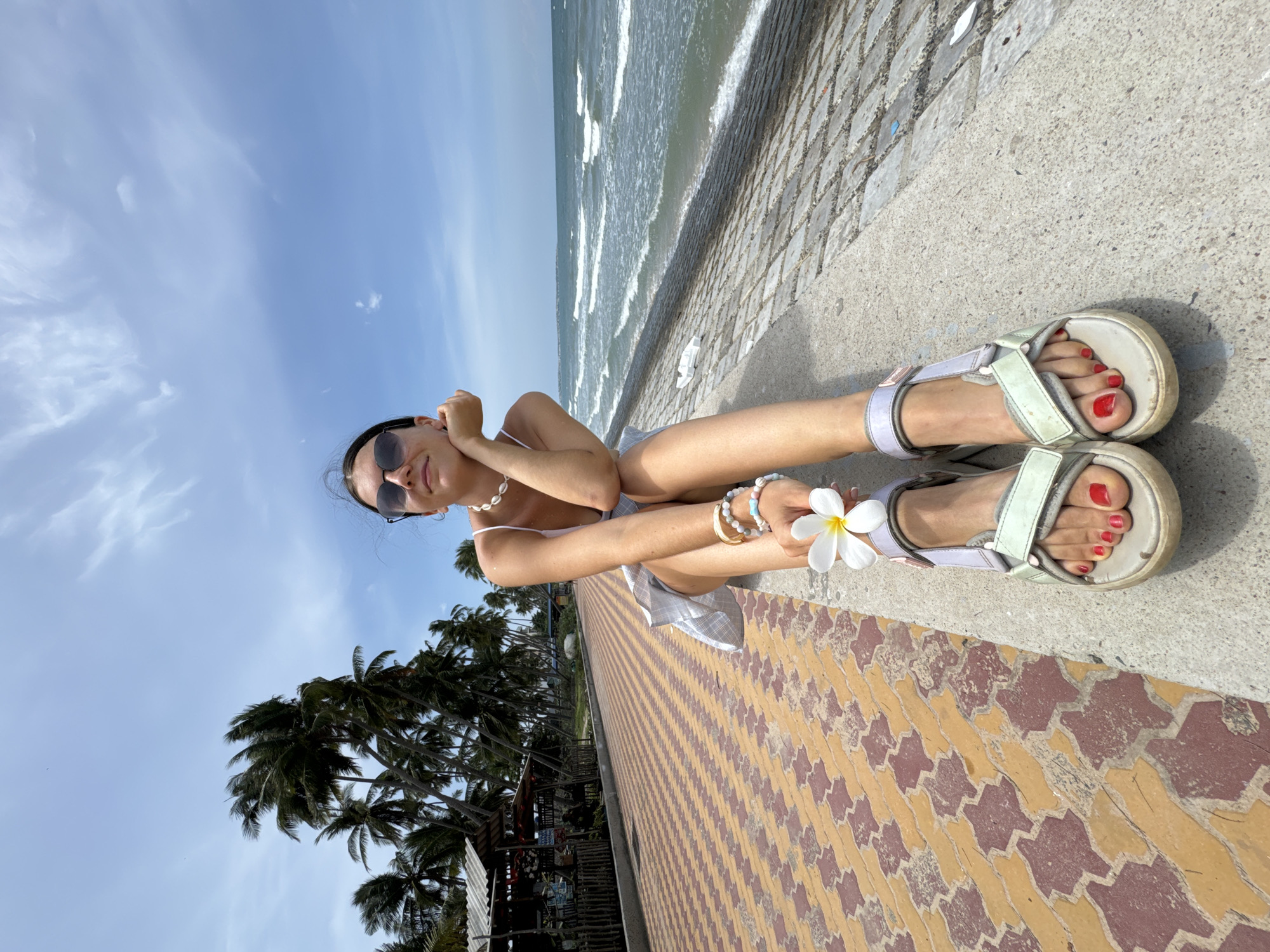 A woman is sitting on a concrete wall by the sea in Mũi Né, Vietnam. She is wearing sunglasses, a white top, and a light-colored skirt. She has bracelets on her left wrist and is holding a white flower in her right hand. Her feet are bare except for sandals, and her toenails are painted red. In the background, palm trees line a paved walkway, and the ocean stretches to the horizon under a blue sky with scattered clouds.