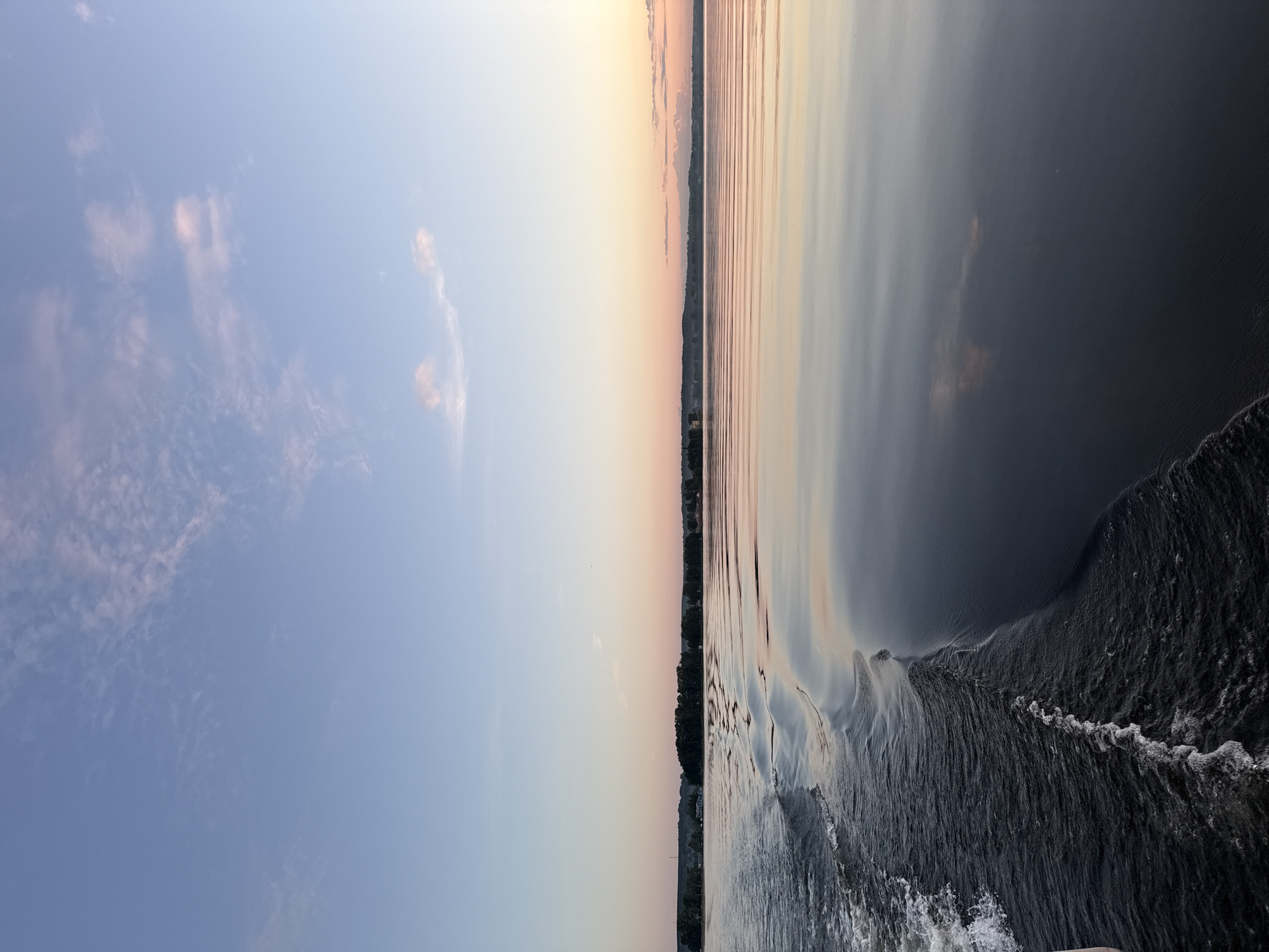 The image displays a vast body of water under a twilight sky, indicative of either sunset or sunrise. The sky in the upper portion is a pale, clear blue, gradually transitioning to soft pink and orange hues at the horizon line. Sparse, wispy clouds, some faintly pink-tinted, are distributed across the sky, particularly in the upper left and near the horizon.
The water surface dominates the lower two-thirds of the frame, reflecting the sky's coloration in shades of deep blue-grey in the foreground, lightening to lighter blues, grays, and subtle pinks further away. A distinct boat wake originates from the lower-left corner, characterized by white aerated foam near its inception, evolving into a series of rhythmic, dark, undulating wave patterns that extend diagonally across the water. The water on the right side of the image is notably calmer, exhibiting smooth reflections.
A narrow, distant landmass forms the horizon, separating the water from the sky. This land appears to be heavily forested with a dense line of dark green trees. Faint, indistinct structures, possibly buildings, are minimally visible within or behind the tree line, predominantly on the left side of the land strip. The perspective suggests the photograph was captured from the aft of a moving vessel. There are no individuals or identifiable objects other than the boat's wake.
This scene is consistent with the Votkinsk Pond (Votkinskoye Vodokhranilishche), a large artificial lake or reservoir in Votkinsk, Russia, known for its expansive waters and wooded shores.
