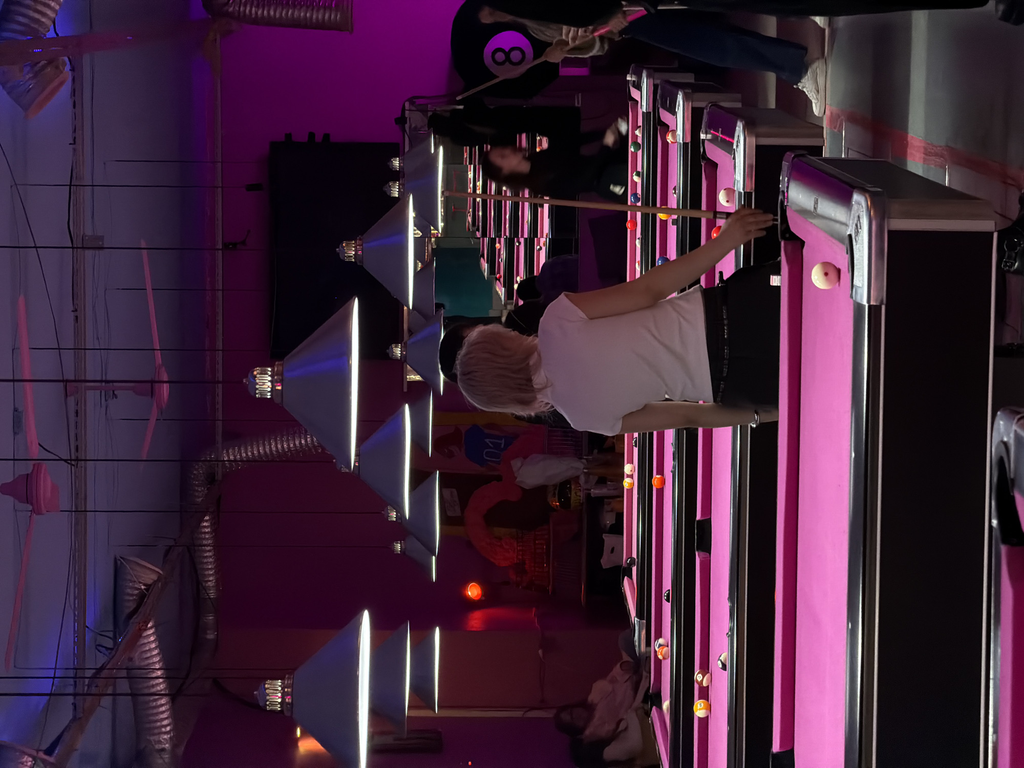 The image shows a billiards hall in Saint Petersburg, Russia, with rows of pink billiards tables under bright pendant lights. Several people are playing billiards, including one woman with blonde hair in a white shirt who is preparing to take a shot. A person wearing a black shirt with an "8" ball design is standing at a table in the background. The environment has a purple hue from the lighting, and ductwork is visible on the ceiling.