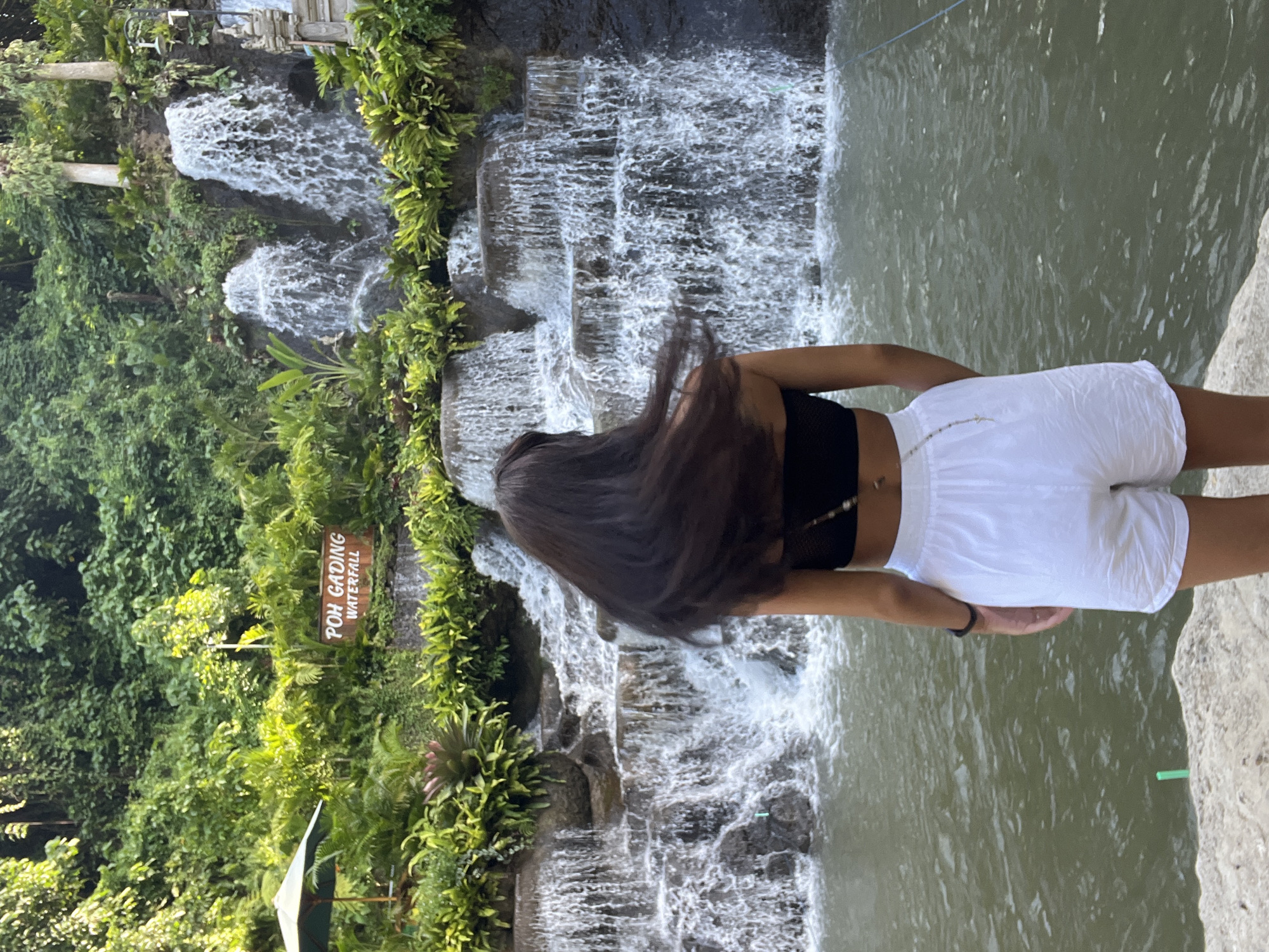 A woman in a black top and white shorts stands at Poh Gading Waterfall in Sukawati, Indonesia. She is positioned on a stone ledge overlooking the waterfall and a pool of water. Her long dark hair is swept forward, obscuring her face. Lush green vegetation and tiered waterfalls form the backdrop. A sign reads "POH GADING WATERFALL".