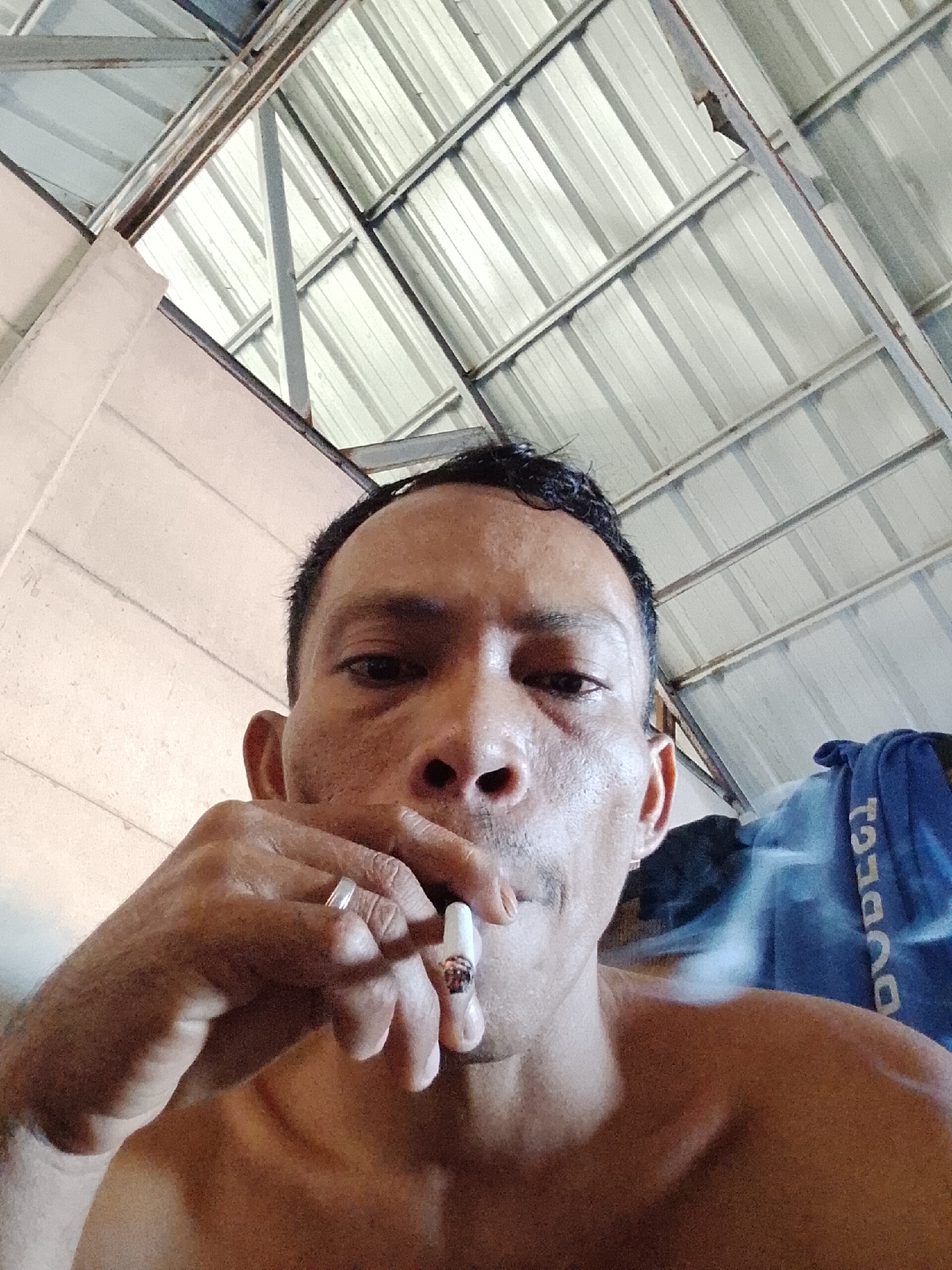 A shirtless man is smoking a cigarette indoors, with the city of Bukit Ketok, Indonesia as the location. He is wearing a ring on his left hand. The interior has a corrugated metal roof. A partially visible blue towel with white text is present in the background. The man's skin is weathered. There is smoke emanating from the cigarette.
