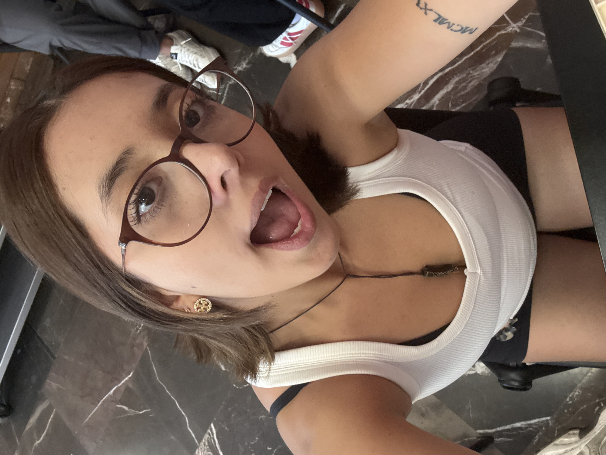 A young woman is looking up into the camera with an open mouth expression. She is wearing glasses and a white tank top. A small gold earring is visible on her left ear. A tattoo is visible on her arm. The background includes a marble-patterned surface. The city of San Andrés Cholula, Mexico, cannot be determined from the available visual information.