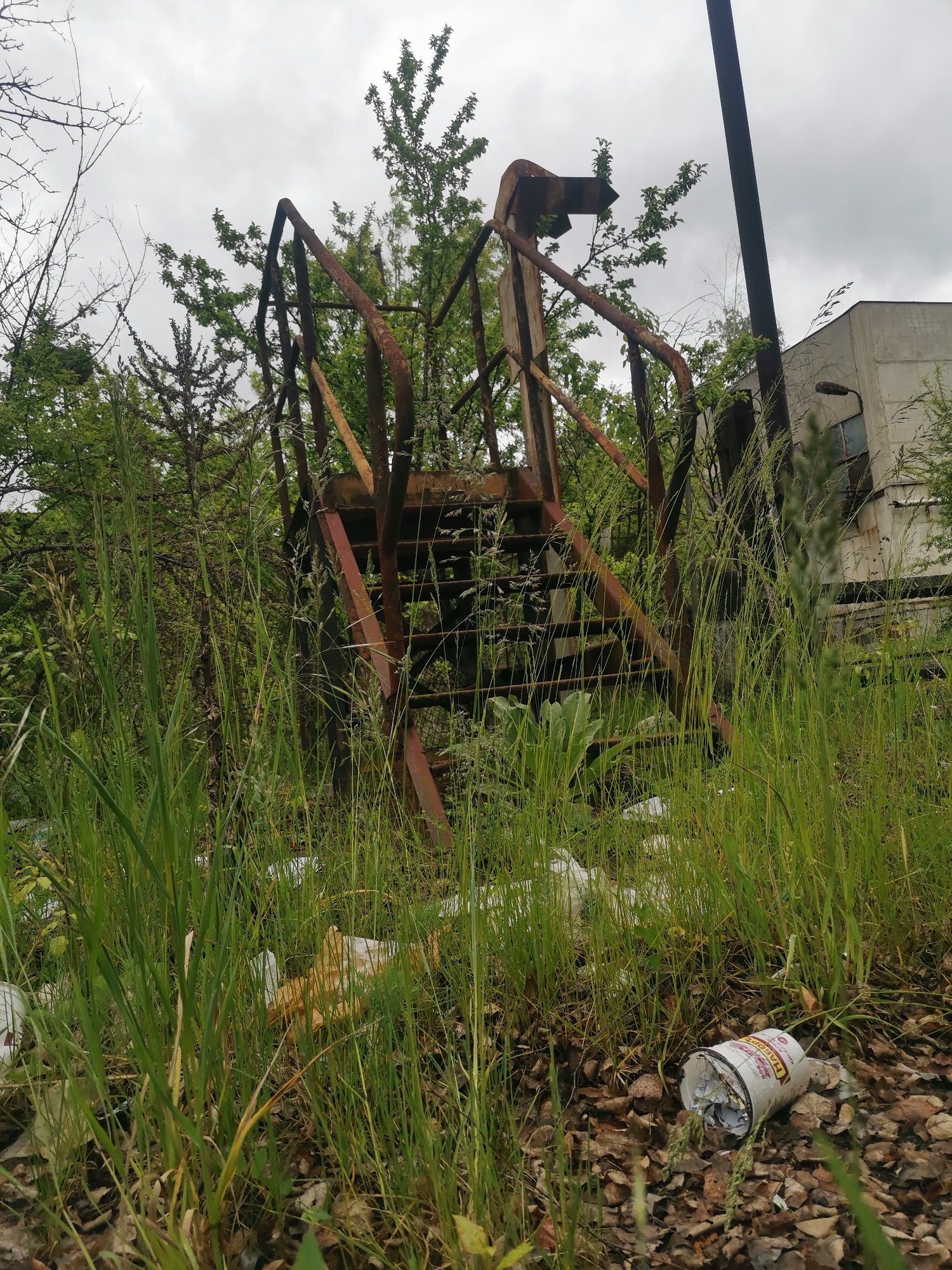 The image shows an overgrown, neglected area in Verinsko, Bulgaria. A rusted, metal staircase stands amidst tall grass and vegetation, indicating disuse and abandonment. Litter, including a discarded paper cup, is scattered on the ground, contributing to the unkempt appearance. A concrete building is partially visible in the background, suggesting an industrial or commercial zone. The sky is overcast, creating a somber atmosphere. There are no visible people or actions. The scene depicts urban decay and environmental neglect.