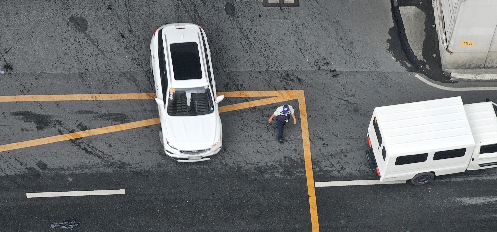An aerial view shows a street scene in Makati, Philippines. Two white vehicles are present: a modern SUV parked on the left side of the road and a utility van parked on the right. A person in a uniform, appearing to be a security guard or similar personnel, is walking on the asphalt between the two vehicles. The ground is wet, suggesting recent rain or it being a damp day. Yellow lines mark parking spots or lane divisions on the road. A portion of a concrete structure with a yellow label reading "ED 15" is visible on the right side of the image. The lighting suggests it is daytime, though the overcast conditions may obscure direct sunlight. The overall scene depicts an ordinary street environment with traffic and pedestrian activity.