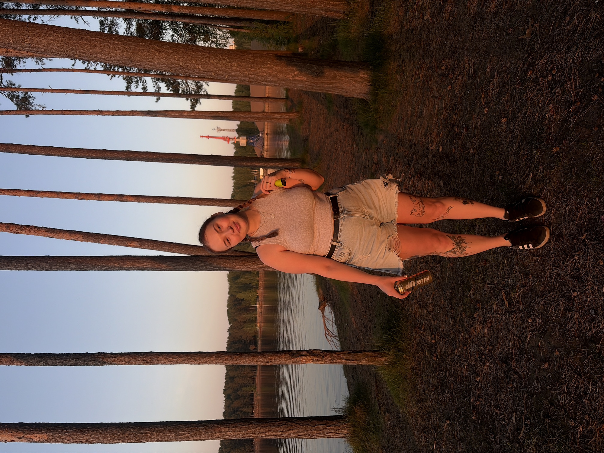 The image shows a woman lying on the ground near a lake and a line of pine trees. The woman is wearing a gray top, denim shorts, a black belt, and black sneakers. She has a braid and is holding a yellow ball in one hand and a can of "Pulse Up" energy drink in the other. Tattoos are visible on her legs and arm. In the background, across the lake, are trees and a tall red and white structure that appears to be an amusement park ride. The ground is covered with pine needles. The location is likely in the city of Chaikovsky, Russia, suggested by the "Pulse Up" can.