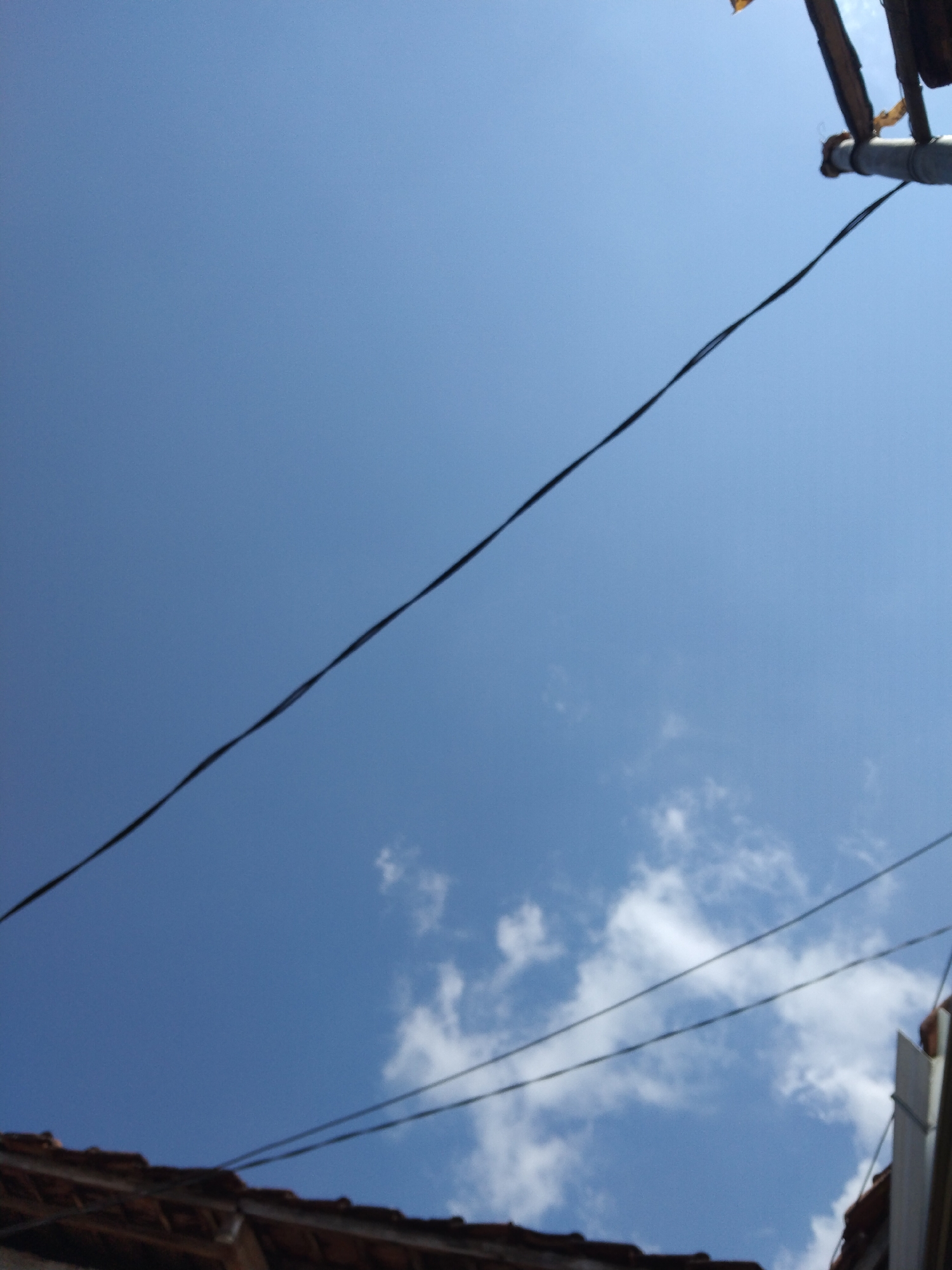 The image depicts a clear blue sky with a few scattered white clouds. Several black electrical wires stretch across the frame, some of them appearing taut while others have a slight sag. In the lower portion of the image, the terracotta tile roof of a building is visible, suggesting an outdoor scene in a built-up area. The perspective is looking upwards, emphasizing the expanse of the sky. This scene is likely from the city of Kelet, Indonesia, given the context.
