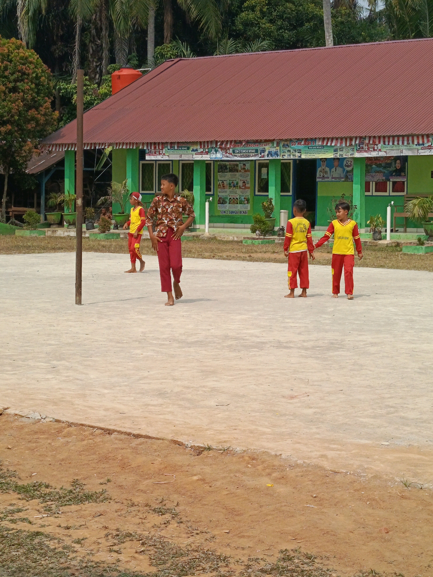 The image depicts an outdoor scene in front of a school building in Megang Sakti IV, Indonesia. A group of four young boys, dressed in school uniforms consisting of yellow long-sleeved shirts and red pants, are present on a concrete ground. One boy, in the foreground and slightly to the right, is wearing a patterned brown and white short-sleeved shirt and red pants. The school building, painted green with a red roof, features several windows and is decorated with posters and banners. A tall wooden pole stands in the foreground, slightly to the left of center. The ground appears to be a mixture of concrete and dirt, with some sparse green vegetation visible in the background and along the edges of the scene. Tall trees with dense foliage are present behind the school building, suggesting a tropical environment. The lighting indicates it is daytime.