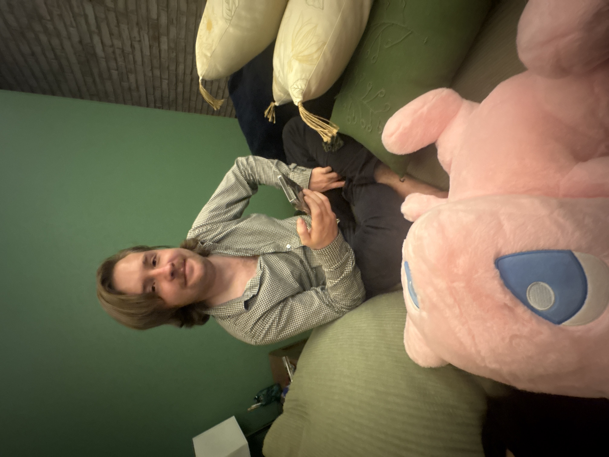 A person is reclining on a sofa surrounded by pillows and a large pink plush toy. The person has light brown hair and is wearing a patterned button-down shirt and dark pants. They are holding a small, rectangular object in their hands. The sofa has multiple pillows of varying colors and textures. In the background, there is a green wall and a textured ceiling. Near the sofa, there appears to be a side table with some items on it.
This information does not describe any location in the city of Huixquilucan de Degollado, Mexico.