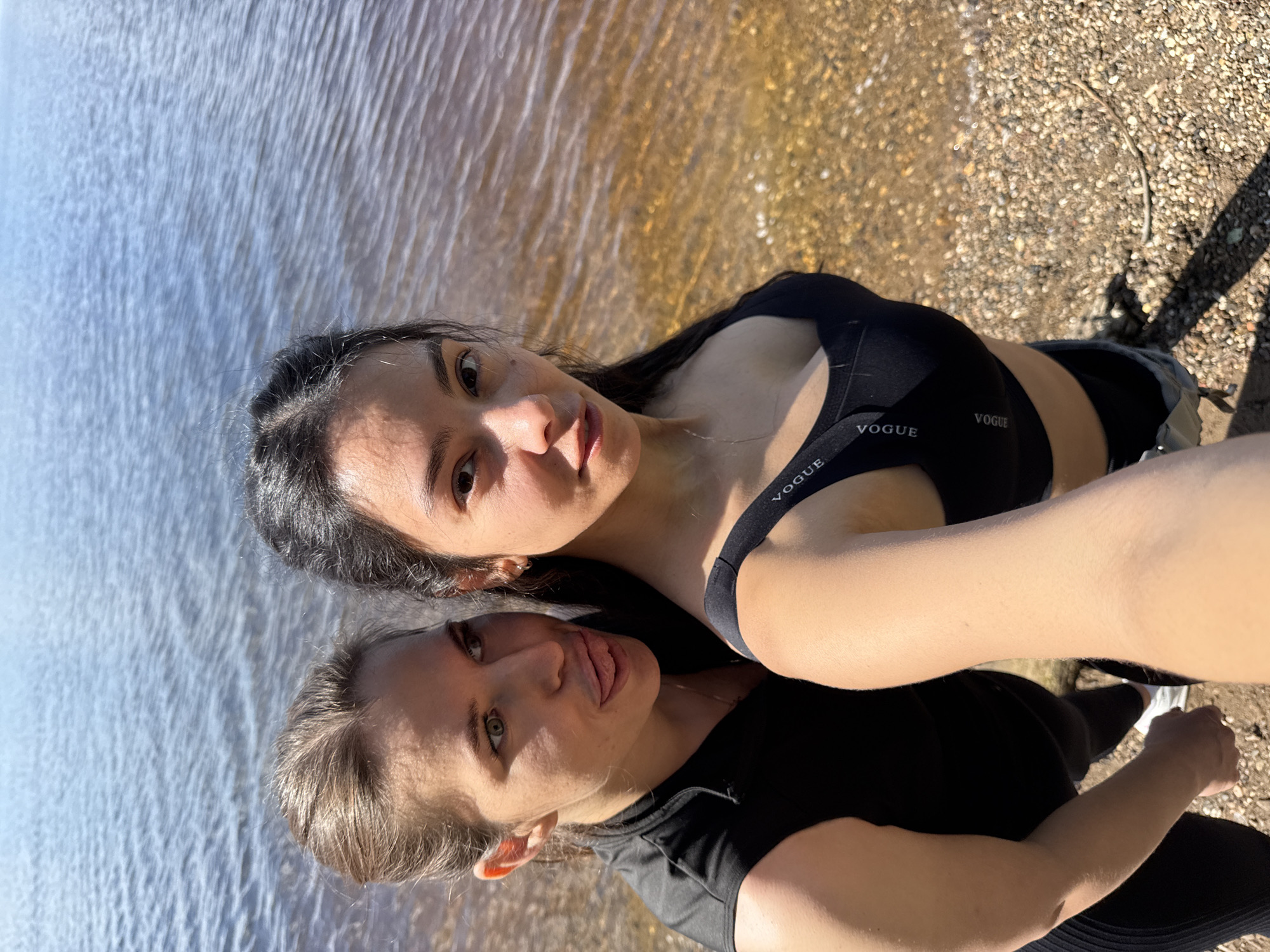 Two women are captured in a selfie at a pebbled shoreline in the city of Chaikovsky, Russia, with a large body of water in the background under clear, sunny conditions. The woman in the foreground, positioned to the right, holds the camera, looking directly forward with a neutral expression. She has dark hair and is wearing a black sports bra with "VOGUE" printed repeatedly on the shoulder straps. The second woman, positioned behind and to the left, has light brown hair pulled back and green eyes. She is looking to the right of the frame, sticking her tongue out in a playful gesture. She wears a black sleeveless athletic top. The water exhibits small ripples, indicating a light breeze, and transitions from blue-grey to a clearer, brownish hue near the shore, revealing small, light-colored pebbles and some scattered larger stones and debris on the ground. Shadows from the subjects are visible on the shore.
