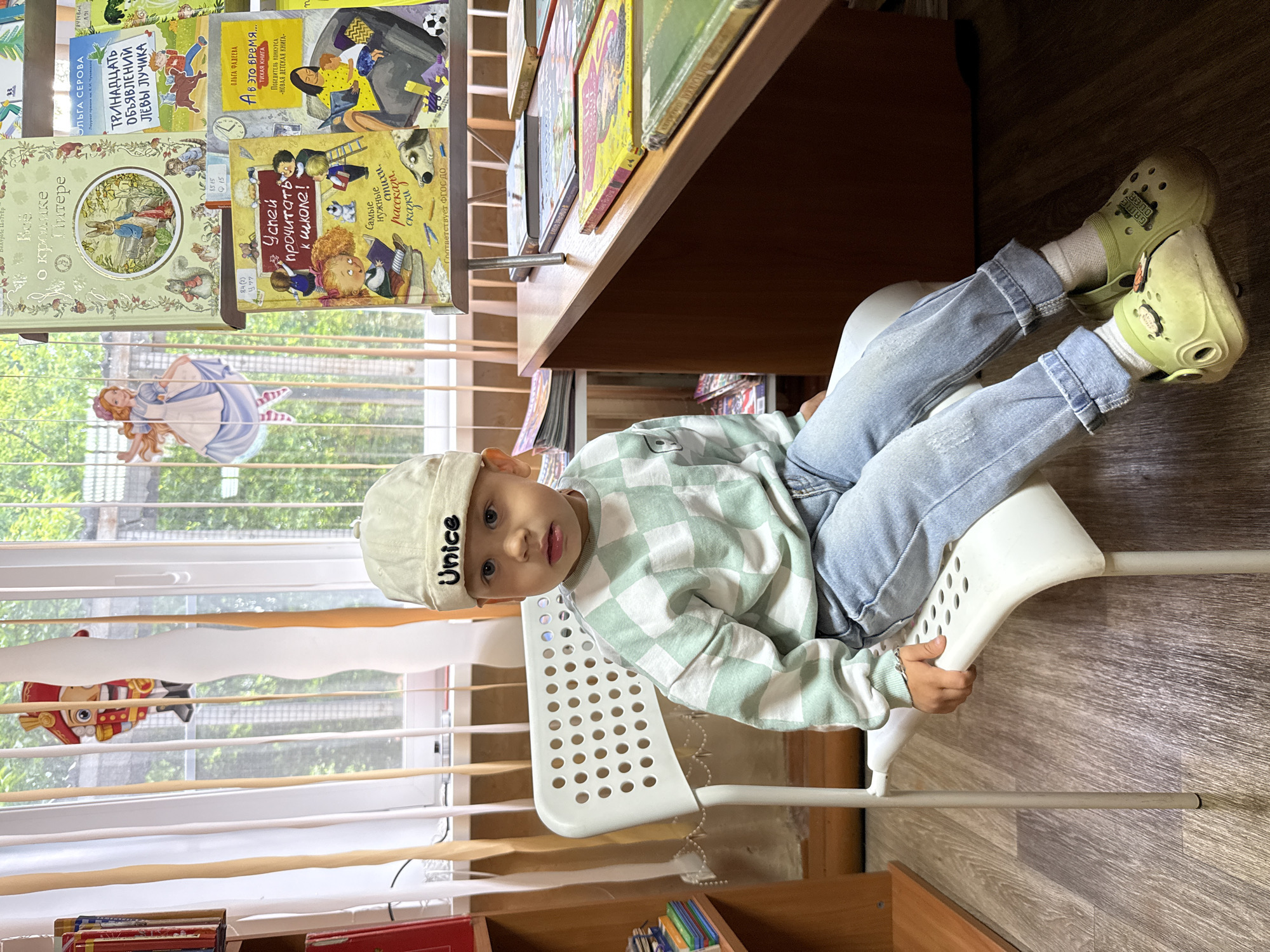 A young child sits on a white chair indoors, looking directly at the camera. The child wears a light-colored cap with "Unice" written on it, a light green and white checkered long-sleeved shirt, light blue jeans, white socks, and lime green Crocs with "GAME OVER" text on them. The setting appears to be a children's library, bookstore, or a reading area, indicated by numerous children's books displayed on shelves.
To the child's left and behind them, a window with blinds is visible, showing green foliage outside, suggesting it is daytime. Decorative cutouts of characters like Alice in Wonderland and a Nutcracker figure are seen hanging or pasted on the window. The floor is wooden.
Several book titles are visible, including: "Всё о кролике Питере" (All about Peter Rabbit), "ОЛЬГА СЕРОВА ТРИНАДЦАТЬ ОБЪЯВЛЕНИЙ ЛЁВЫ ЛУЧИКА" (Olga Serova Thirteen Announcements of Lyova Luchik), "Успей прочитать к школе!" (Hurry up and read for school!), and "ОЛЬГА ФАДЕЕВА А в это время... - ТИХАЯ КНИГА" (Olga Fadeeva Meanwhile... - QUIET BOOK), which is also labeled "ПОБЕДИТЕЛЬ КОНКУРСА - НОВАЯ ДЕТСКАЯ КНИГА" (WINNER OF THE COMPETITION - NEW CHILDREN'S BOOK). Another book is titled "Самые нужные стихи, рассказы, сказки" (Most essential poems, stories, fairy tales) and states "соответствует ФГОС ДО" (corresponds to Federal State Educational Standard for Preschool Education). The child's expression is one of calm curiosity. The location context is Rusanovo, Russia.