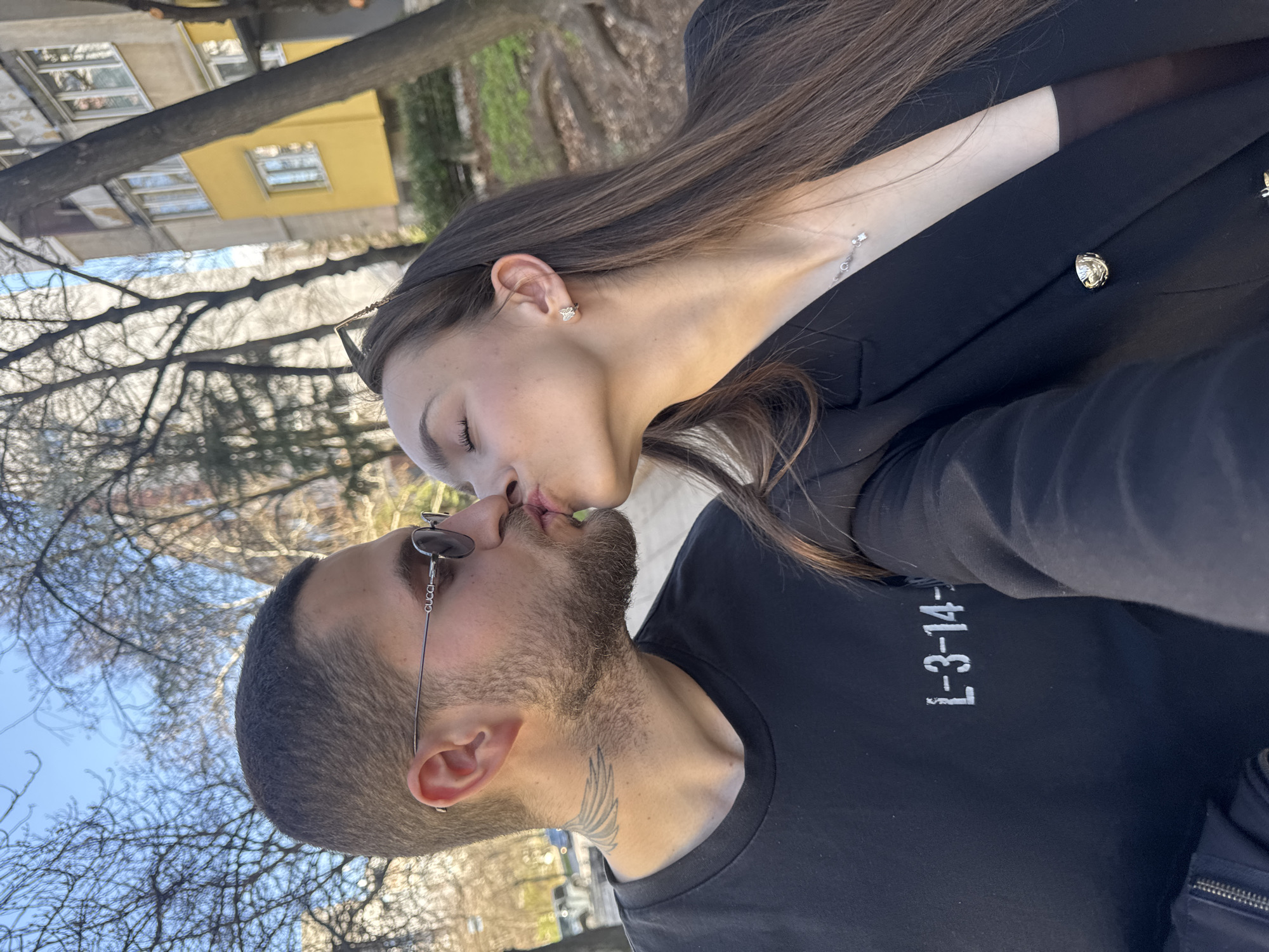 A couple kisses outside on a clear day. The woman has long brown hair and wears a black blazer, a delicate necklace, and diamond-like earrings. The man has a short haircut, a beard, and a tattoo on his neck. He wears sunglasses and a black t-shirt with "L-3-14-" printed in white on the front. The scene is set in a park in Sofia, Bulgaria, with leafless trees and a yellow building in the background.