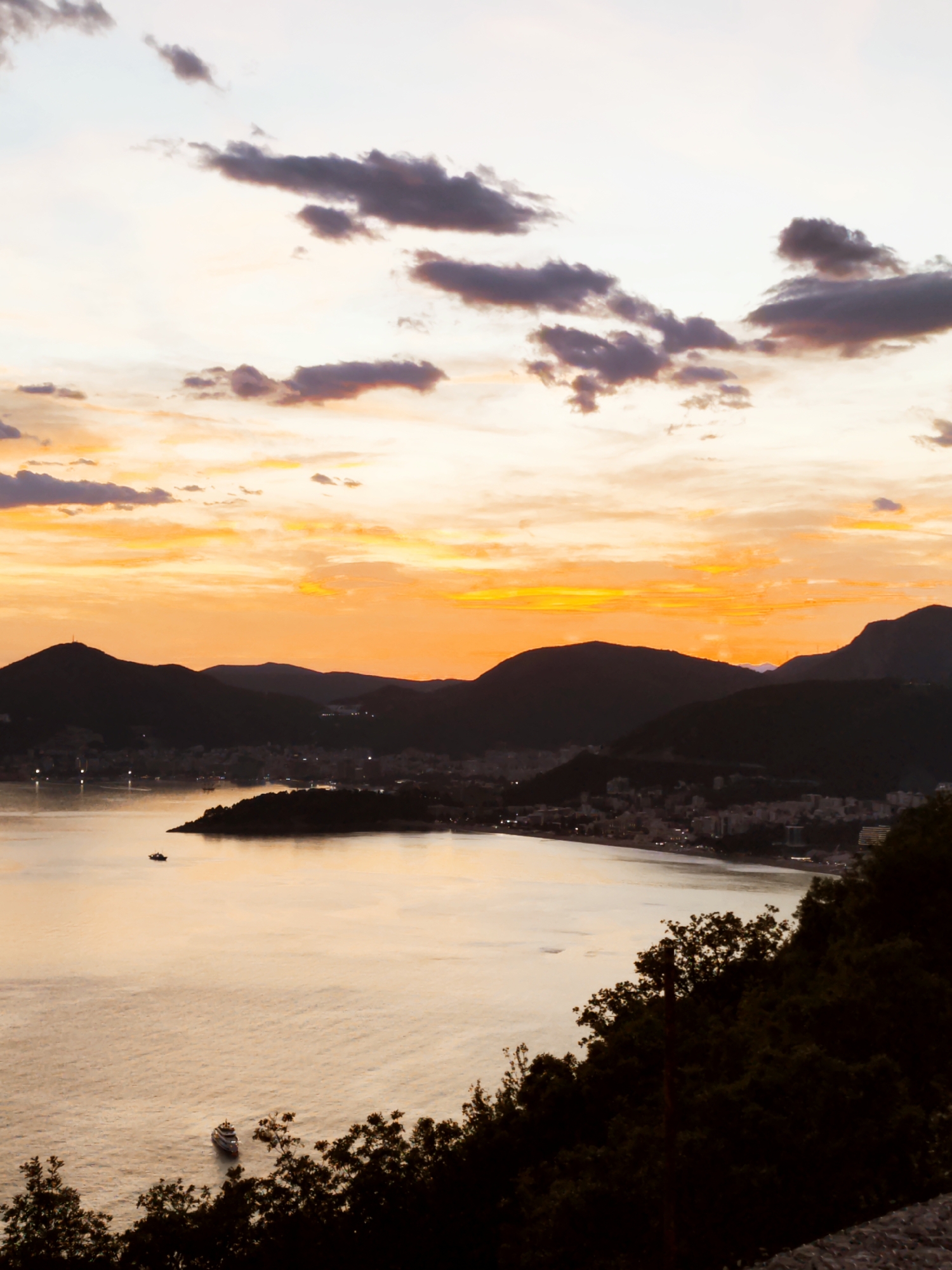 The image shows a coastal city at sunset. The sky is filled with orange and yellow hues, with dark clouds scattered across the top. Below the sky, there are silhouettes of mountains. In front of the mountains, there is a city with lights visible. The water reflects the colors of the sky. There are two boats visible on the water. The foreground of the image contains dark foliage. The location is possibly coastal.