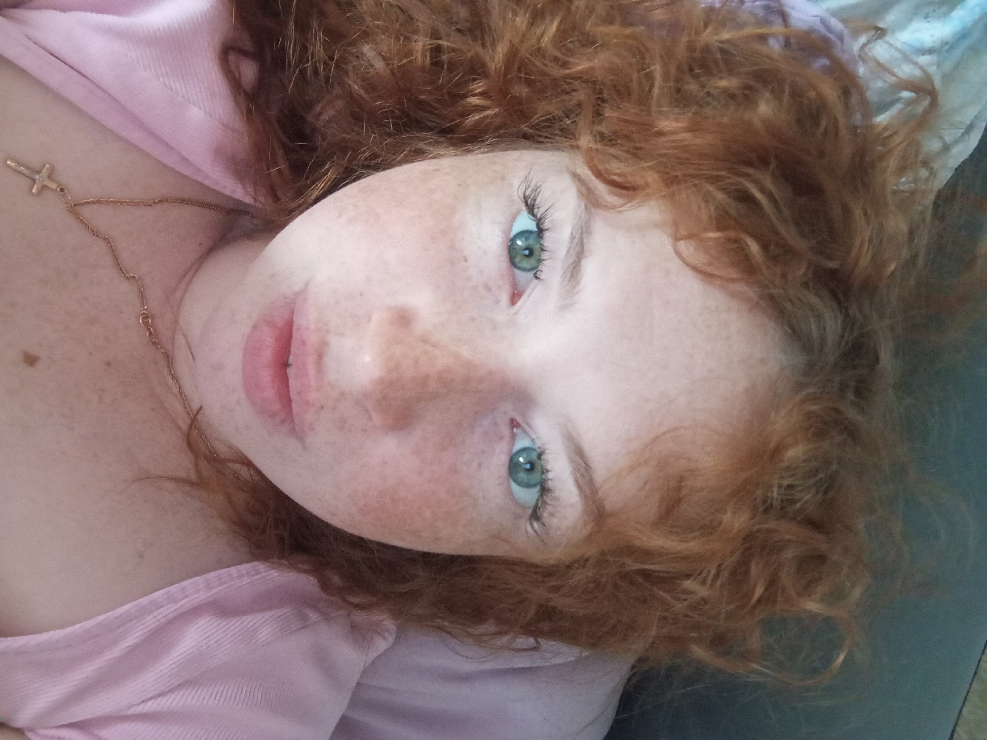 A selfie-style photograph shows a person with red, curly hair and freckled skin. The person has green eyes and is wearing a light pink top. A gold necklace with a cross pendant is visible on their chest. The background appears to be a bed or similar soft surface, with a bluish hue in the upper right corner. The location in Yurgamysh, Russia is not discernible from the image.