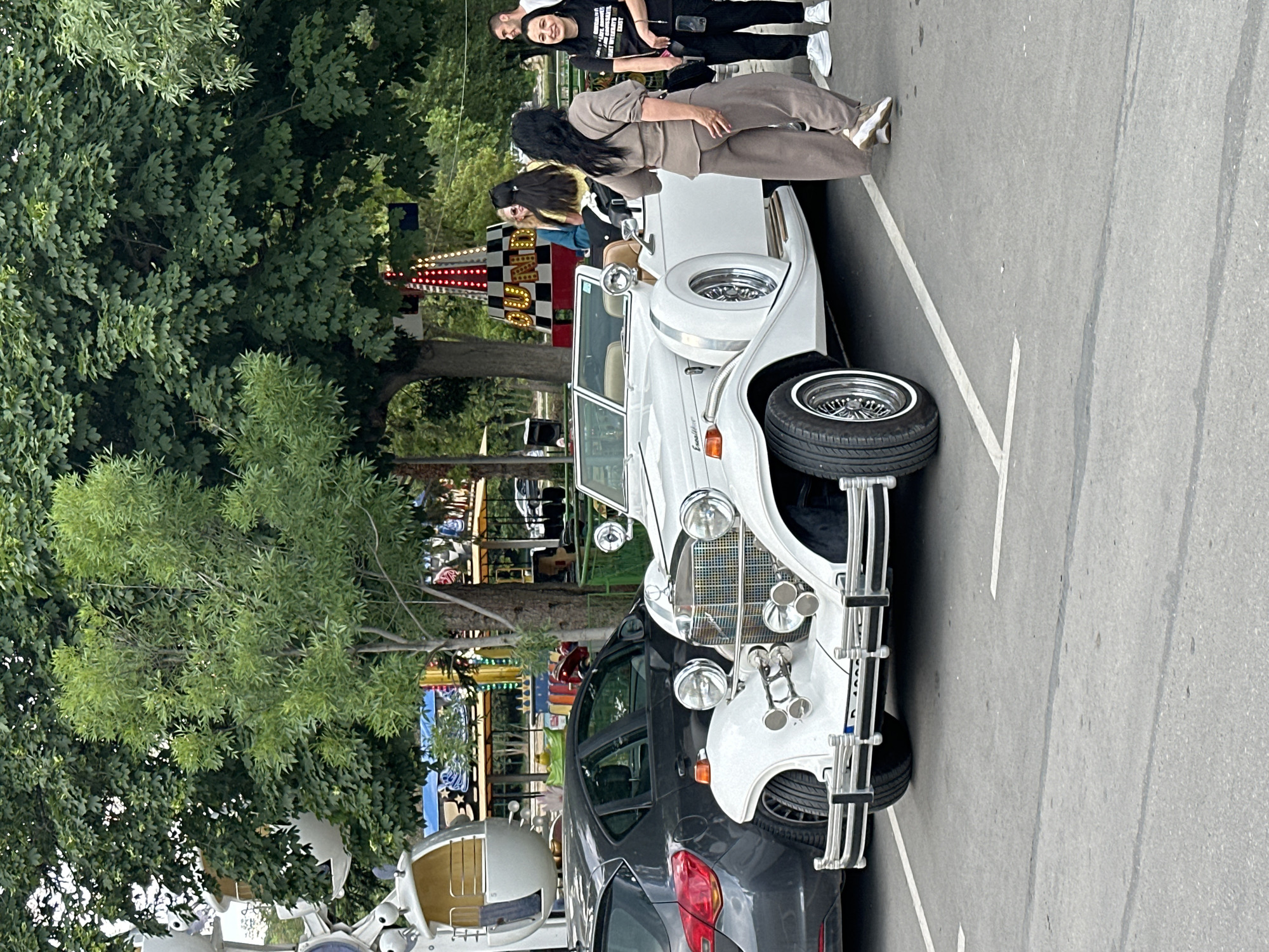 A white vintage car is parked in a lot on its side, appearing as if it is on display for people to look at. Two women stand next to the car and appear to be conversing while another two stand a short distance away behind the car, also conversing. Trees are visible in the background, along with some carnival attractions. Based on the carnival attractions visible in the background, this scene likely takes place in Varna, Bulgaria at the Sea Garden, as it hosts an amusement park.