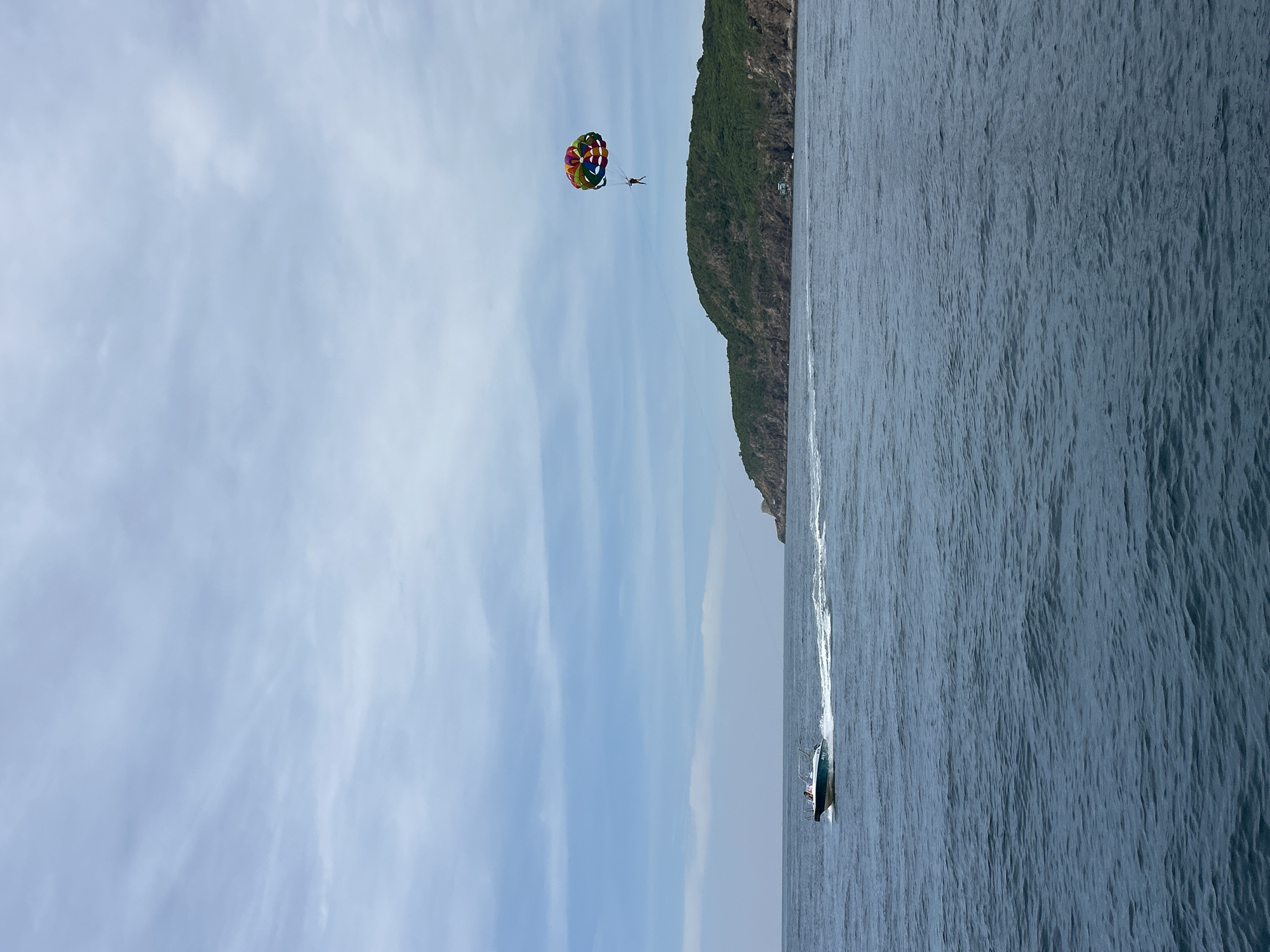 This image captures a coastal scene in Nha Trang, Vietnam. A speed boat, white with a blue trim, is moving from left to right across the deep blue sea, leaving a white wake. The boat appears to have at least two occupants. In the sky to the right of the boat, a person is parasailing, attached to a colorful parachute. Behind the water and to the right, a lush green, hilly island rises from the sea, its rocky shores visible. The sky is a pale blue, with scattered white clouds. The overall impression is one of leisure and recreation on a slightly overcast day.