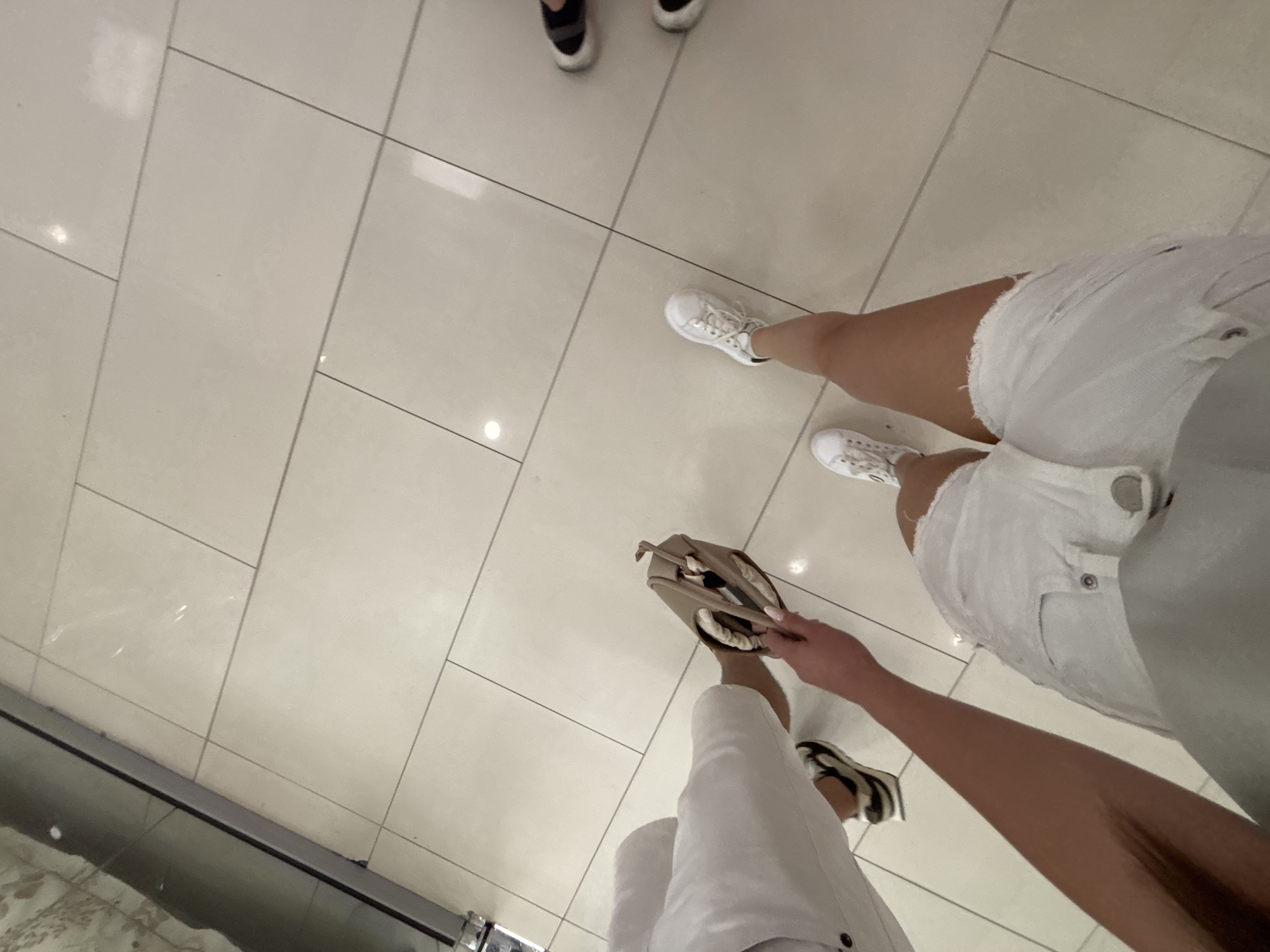 The image presents a low-angle, downward-looking perspective of a tiled floor and the lower extremities of at least three individuals, located in Plovdiv, Bulgaria. The floor is composed of large, light beige or off-white rectangular tiles with dark gray grout lines, exhibiting subtle reflections of overhead lighting. Along the upper-left edge, a dark, vertical metallic or glass fixture is partially visible.
In the immediate foreground, an individual wears white denim shorts with frayed hems and metallic buttons/rivets, paired with white sneakers. This person's left arm extends into the frame, holding an open, light brown bag or clutch. The hand holding the bag has light-colored nail polish, and a striped item is partially visible inside the bag. To the left and slightly behind, another person's lower leg and foot are visible, clad in white trousers and shoes with a distinct brown and black pattern. Further to the right and in the background, a third individual stands, identified by dark-colored shoes with white soles. The scene suggests an indoor public area.