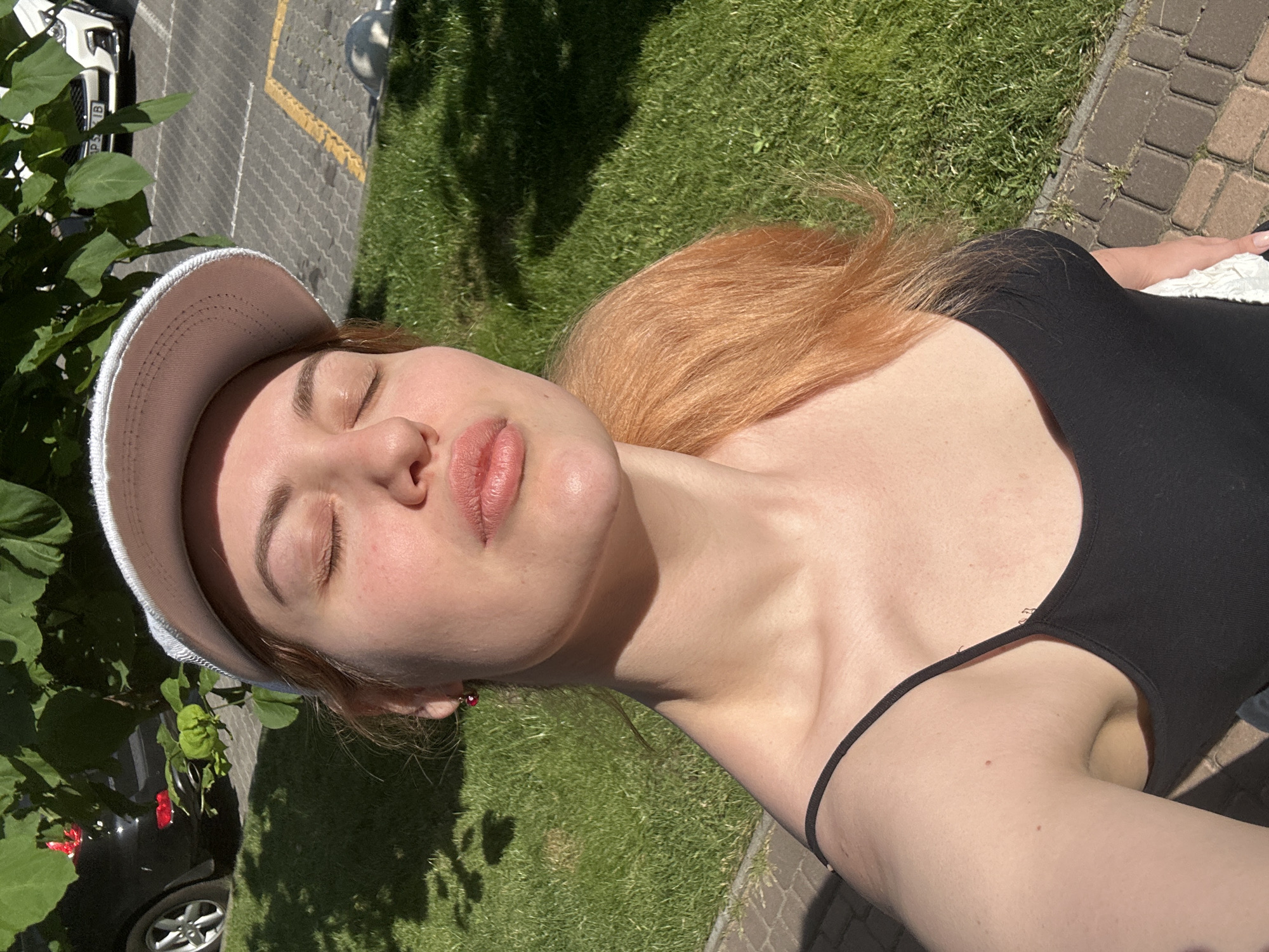 A young woman with fair skin and auburn hair, wearing a black tank top and a white baseball cap, takes a selfie outdoors. She is outdoors in Odesa, Ukraine, with trees, grass, and a paved area visible. The woman's eyes are closed, and her lips are pursed. A black car is parked in the background, along with a white SUV and what appears to be a parking lot. The sun is casting shadows, indicating it is daytime.