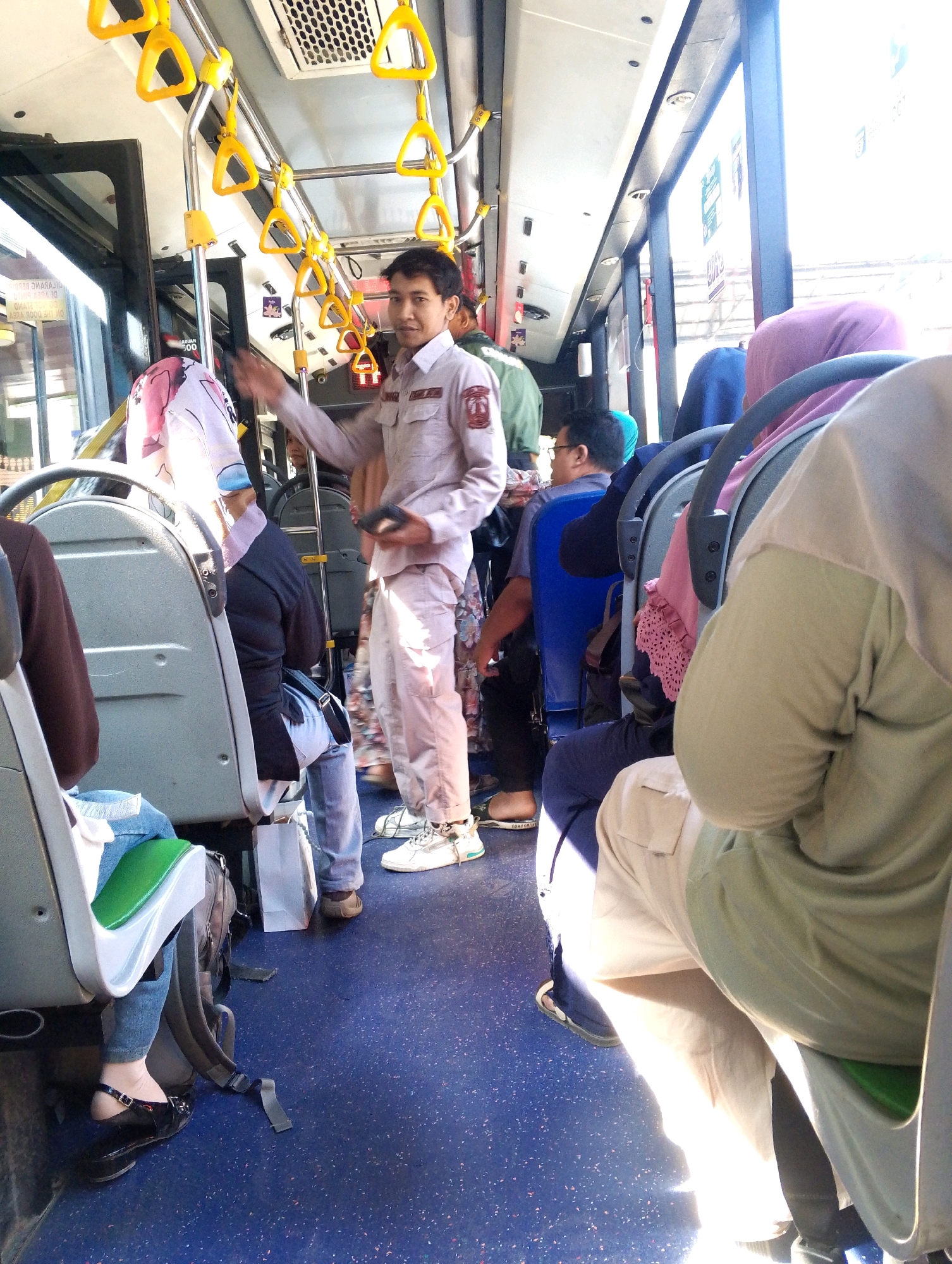 The image captures the interior of a bus in what appears to be a city like Medan, Indonesia, on a bright day with strong sunlight streaming through the windows. In the foreground, several passengers are seated. A woman on the left is wearing a brown long-sleeved top and blue jeans. Next to her, another passenger is partially visible. In the middle of the frame, a male bus attendant, dressed in a light-colored uniform with a badge on his chest and matching trousers, is standing and appears to be interacting with a passenger or making an announcement, holding a dark object in his right hand. He has short dark hair and a light complexion. Behind him, other passengers, some wearing headscarves, are seated. The bus aisle is visible, with blue flooring and numerous yellow handgrips mounted on silver poles overhead. The overall scene depicts a typical commute on a public bus.