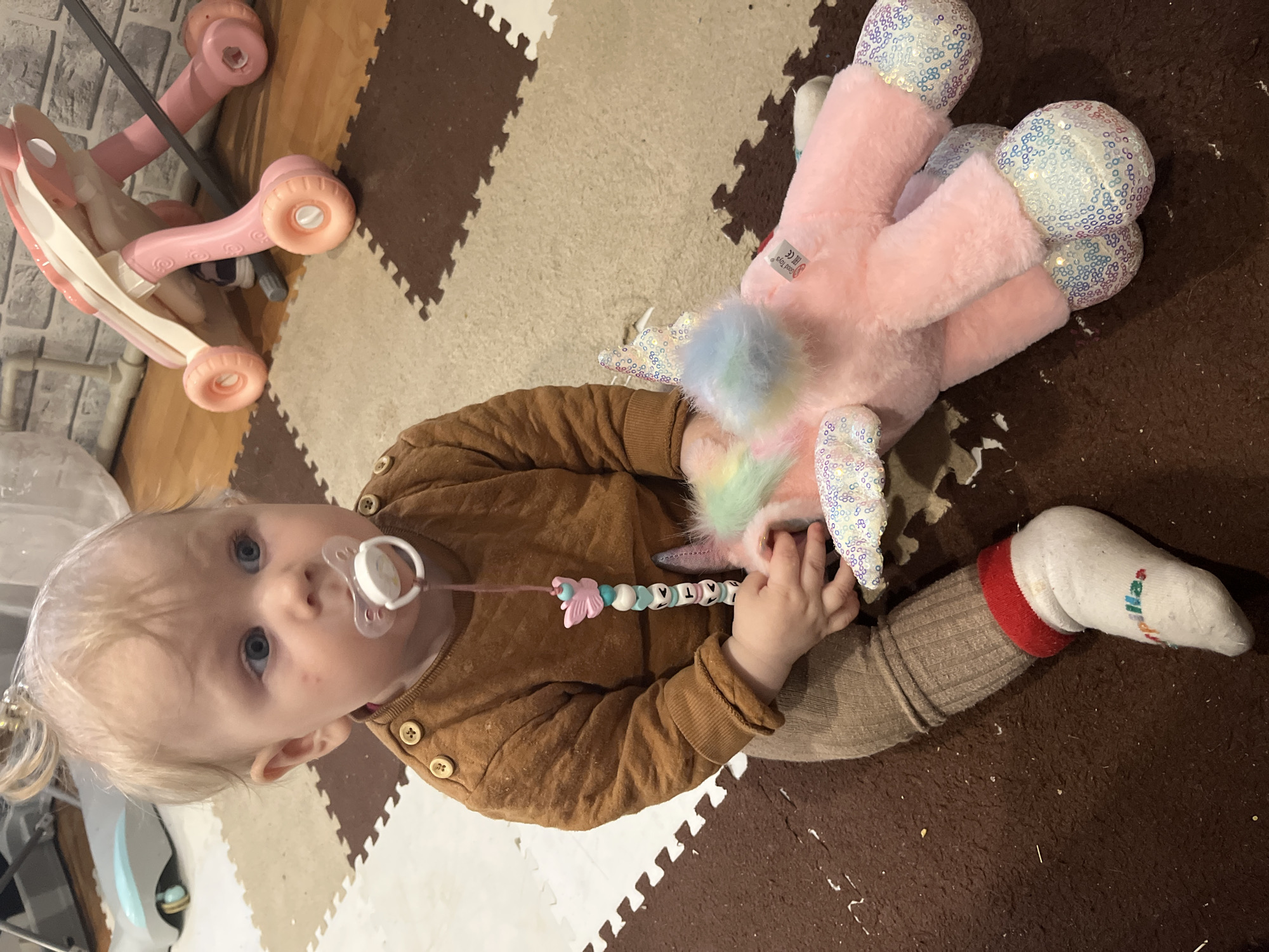 An overhead shot shows a baby or toddler sitting on a floor covered with brown and cream interlocking foam playmats, likely indoors in Vesyoloye, Russia. The infant has light blonde hair, styled in a small ponytail, and blue eyes, looking directly at the camera with a neutral expression. A pacifier is in their mouth, attached to a beaded clip with a pink butterfly charm and visible letters "A" and "T", possibly spelling out a name like "ANASTASIA".
The baby wears a brown quilted long-sleeved top with decorative buttons, brown ribbed leggings, and white socks with red cuffs, one of which has "Stella" partially visible in colorful text. They are holding a large, soft pink plush toy, which appears to be a unicorn or pegasus. This toy features sparkling sequined accents on its hooves, horn, ears, and wings, along with a fluffy, rainbow-colored mane or tail. A small white tag on the plush toy shows "Cool Toys®" and "EAC CE" marks. In the blurred background, against a grey brick-patterned wall, sits a pink child's riding toy with wheels. The scene captures a moment of the child's indoor playtime.