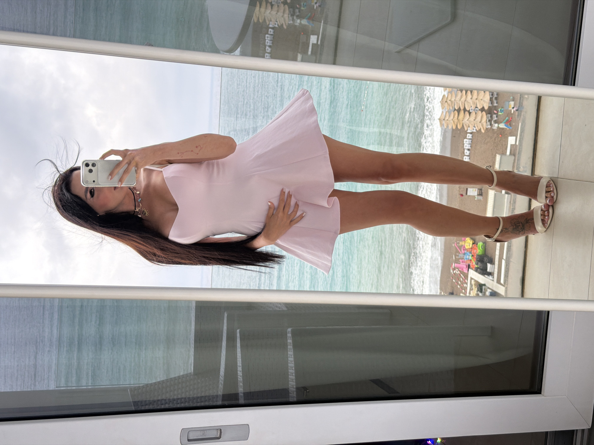 A person is captured in a vertical mirror/reflection selfie, showing from the mid-torso down, wearing a light pink, short-sleeved dress and white open-toed sandals with ankle straps. They hold a silver smartphone to take the picture, largely obscuring their face, with long dark hair visible, some strands appearing wind-swept. A subtle, reddish-brown tattoo resembling constellations or abstract lines is visible on the person's upper left arm, and a floral tattoo adorns their right ankle. Multiple necklaces, including one with a sun-like charm, are visible around their neck. The person's left hand rests casually on their stomach/hip.
The setting is a balcony with white railings and a glass barrier, overlooking a scenic coastline. Under an overcast sky, the calm, turquoise sea stretches to the horizon. Below the balcony, a sandy beach is visible, lined with neat rows of straw beach umbrellas and white sun loungers. Further along the coastline, colorful structures, possibly children's play equipment or small beachside establishments, can be seen. The scene depicts a daytime coastal resort environment in Boreti, Montenegro. No explicit text is visible in the image.