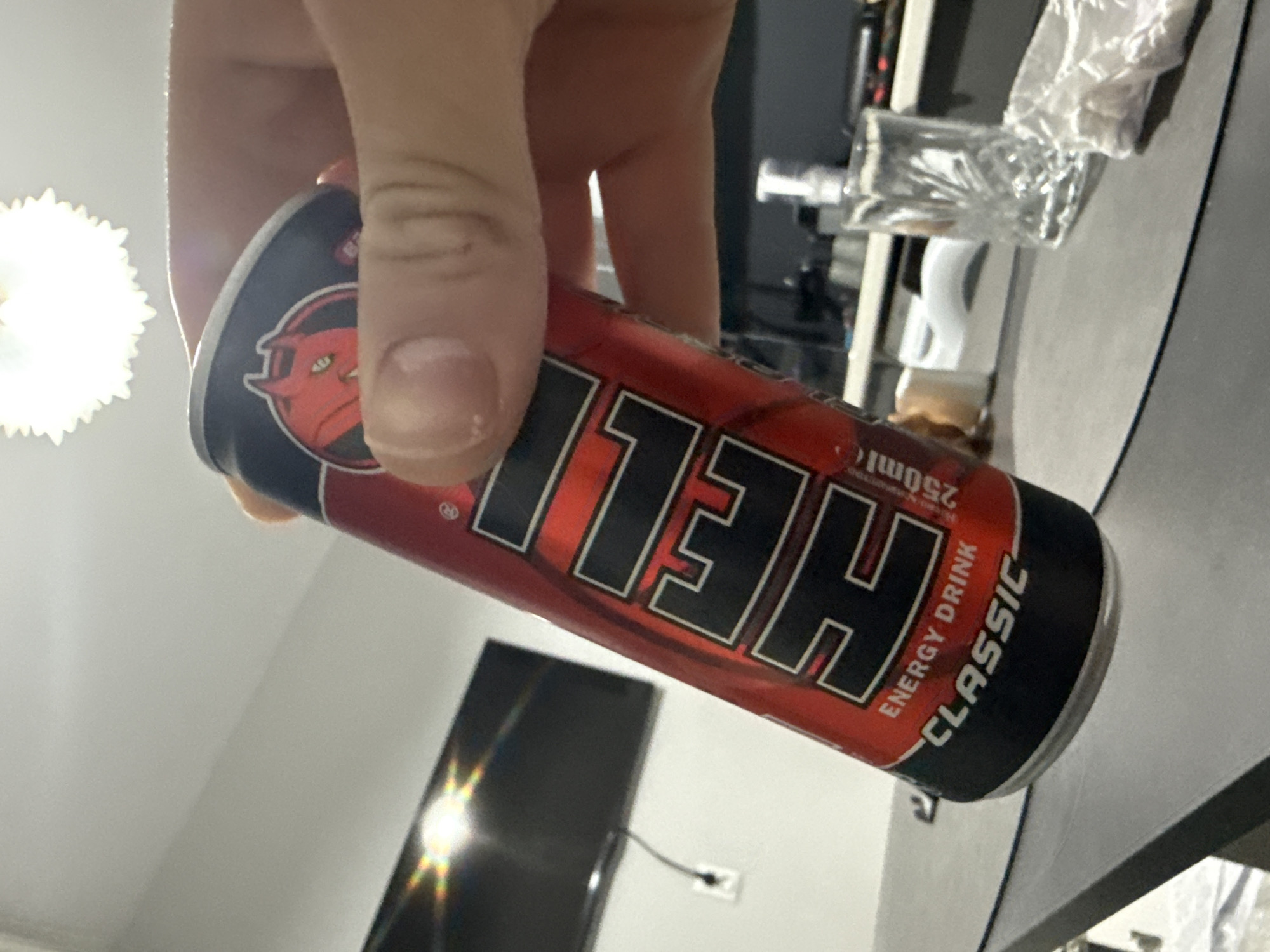 A person is holding a can of "HELL CLASSIC" energy drink in their hand. The can is red and black, with the word "HELL" in large, bold letters. A cartoon devil's head is printed on the top of the can. In the background, there is a partially visible television, a light fixture, and a countertop. The scene is likely indoors, but no specific location in Stara Zagora, Bulgaria can be identified.