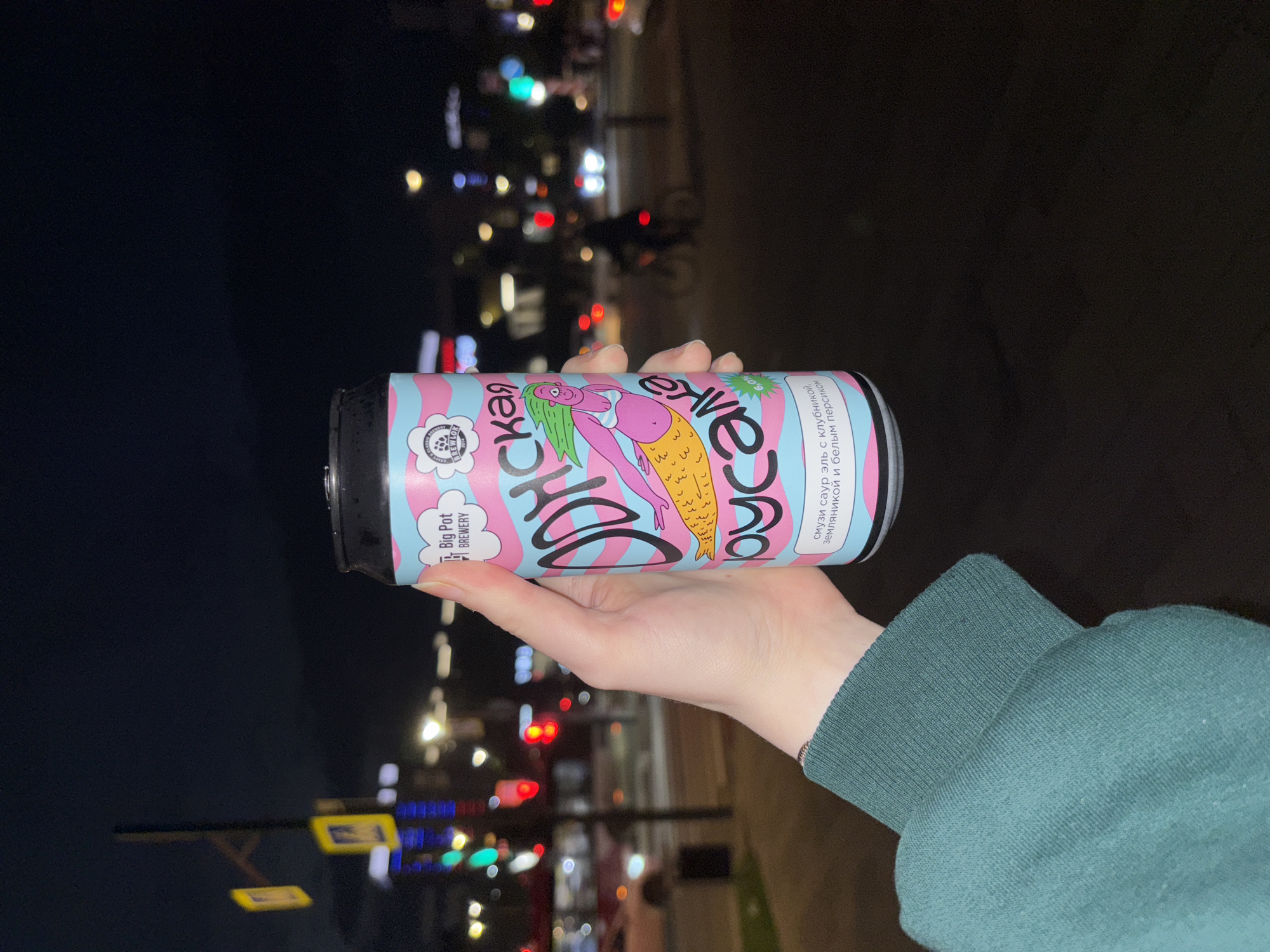 A person's hand, wearing a teal green long-sleeved shirt, holds a can of beer with a colorful, cartoonish design featuring a mermaid. The can has pink and blue wavy stripes. The text on the can is in Russian and reads "ОДУНСКАЯ РУСАЛКА" with "Big Pot BREWERY" printed above it. Below the mermaid illustration, there is smaller text indicating the flavor: "смузи саур эль с клубникой, земляникой и белым персиком" (smoothie sour ale with strawberry, wild strawberry and white peach). The can also displays "6.0%" and the "BREWLOG 2014" logo. The background is out of focus and appears to be an urban setting at night. Streetlights, traffic lights (red and green), and blurred car lights are visible, suggesting a street or intersection. The overall atmosphere is dimly lit, characteristic of a city at night.