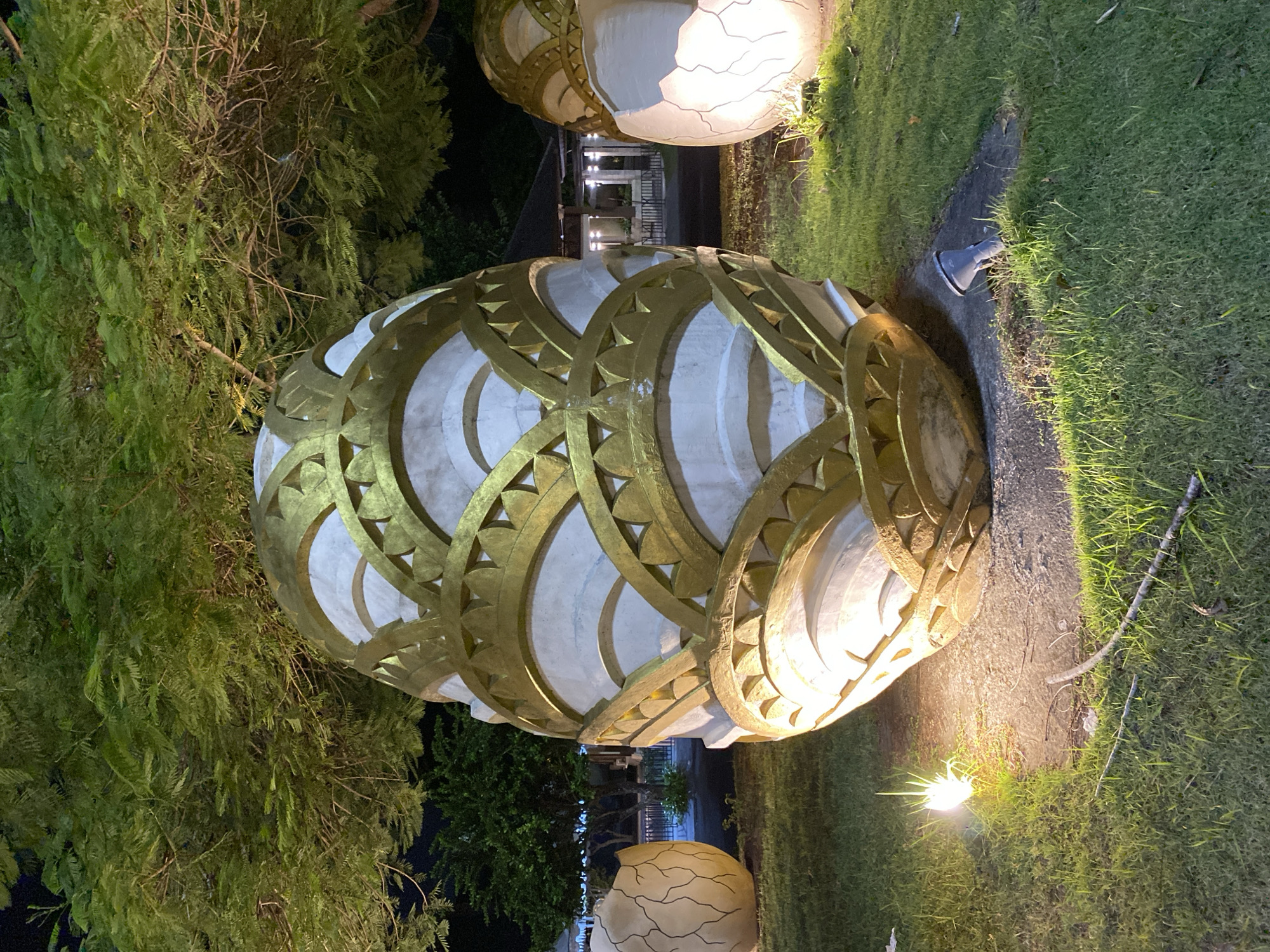 In Kuta Selatan, Indonesia, at night, two large decorative egg sculptures are displayed outdoors on a grassy area. The sculpture in the foreground is elaborately decorated with white and gold patterns that resemble scales or overlapping arcs. It is illuminated by a small spotlight at its base. To the right, another similar egg sculpture is partially visible; it appears to be cracked open and illuminated from within. Behind the sculptures, lush green trees with dense foliage are visible, with their branches extending over the scene. In the background, a dimly lit building with a thatched roof and outdoor seating is visible, suggesting a restaurant or recreational area. The ground is covered with green grass, and some scattered twigs and leaves are present. The overall scene suggests an outdoor decorative installation, possibly in a park or resort setting.