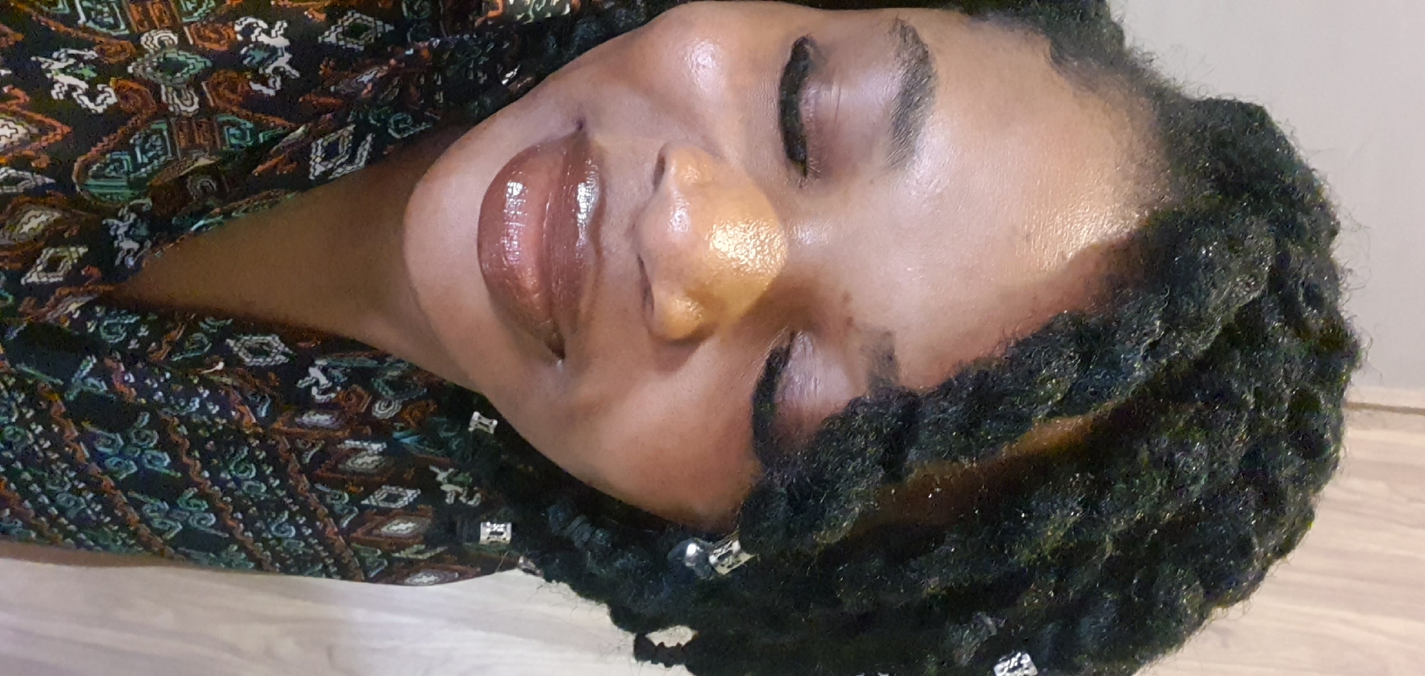The image is a close-up, portrait-style photograph of a woman with dark, braided hair styled into locs, adorned with silver beads. She has closed eyes and a slight smile, with full lips colored in a glossy, brown-toned lipstick. Her skin appears luminous, suggesting makeup. She is wearing a patterned top with geometric designs in various colors, including black, orange, green, and white. The background is a plain, light-colored wall, possibly wood-paneled, and a hint of a beige wall. The image appears to be taken indoors. The specific location of Jos, Nigeria, cannot be determined from the visual information provided in the image.