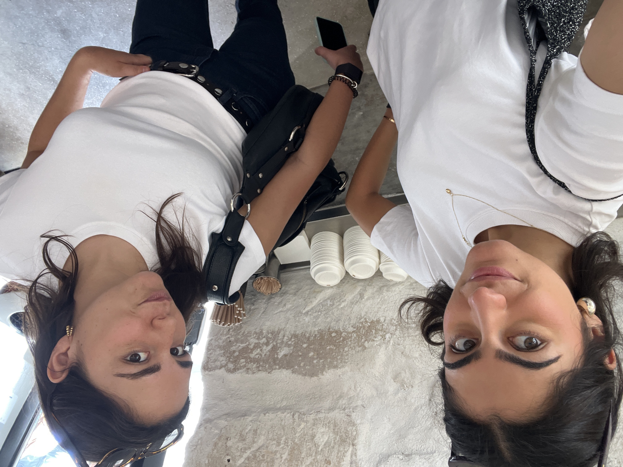 Two women are seen in an upward-angled shot, likely a selfie. Both women wear white T-shirts. The woman on the left wears black pants with a thick belt and a black handbag. She holds a smartphone in her hand and wears a black bracelet. A row of disposable coffee cups are between them. The setting seems to be indoors with a textured white wall. Based on the background architectural style, the location may be Paris, France.