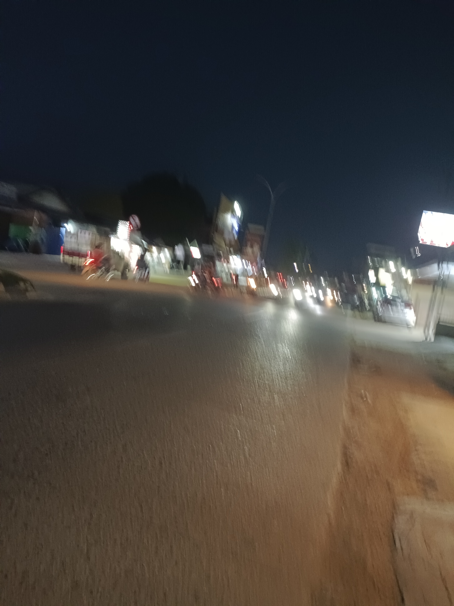 The image captures a blurred, nighttime scene on a street in Teluk Lingga, Indonesia. A road surface, appearing wet and reflective, dominates the foreground. In the mid-ground and background, various out-of-focus elements suggest a street with buildings and moving vehicles. Lights from shops and vehicle headlights create streaks of illumination. Several motorcycles are visible, with some appearing to be in motion. The overall impression is a busy, urban environment at night, characterized by low light and motion blur.