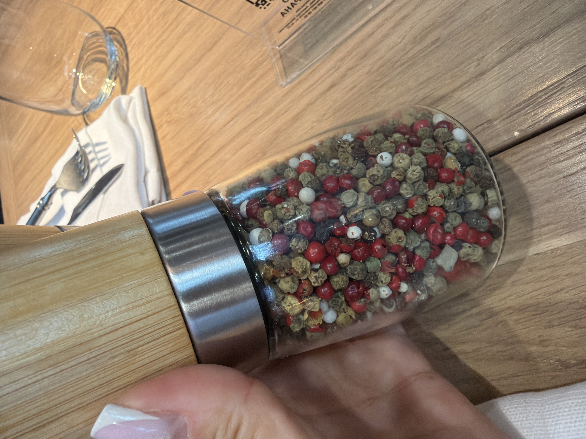 A close-up shot shows a hand holding a peppercorn grinder filled with a mix of red, white, and green peppercorns. The grinder has a bamboo top and a stainless steel collar. The background reveals a wooden table with a white napkin, a fork, a knife, and a glass. A transparent plastic stand with some text, likely a menu or a sign, is also visible on the table. The scene appears to be set in a restaurant or dining establishment in Khimki, Russia.