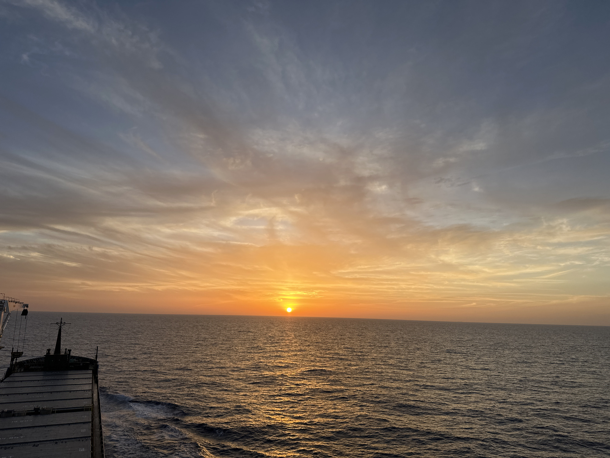A view of the sun setting over the ocean from the deck of a ship. The sun is a bright orange orb on the horizon, reflecting off the water. The sky is filled with streaky clouds, with colors ranging from blue and gray to orange and yellow. The ship's deck and part of its structure are visible in the lower left corner. The location is the ocean.