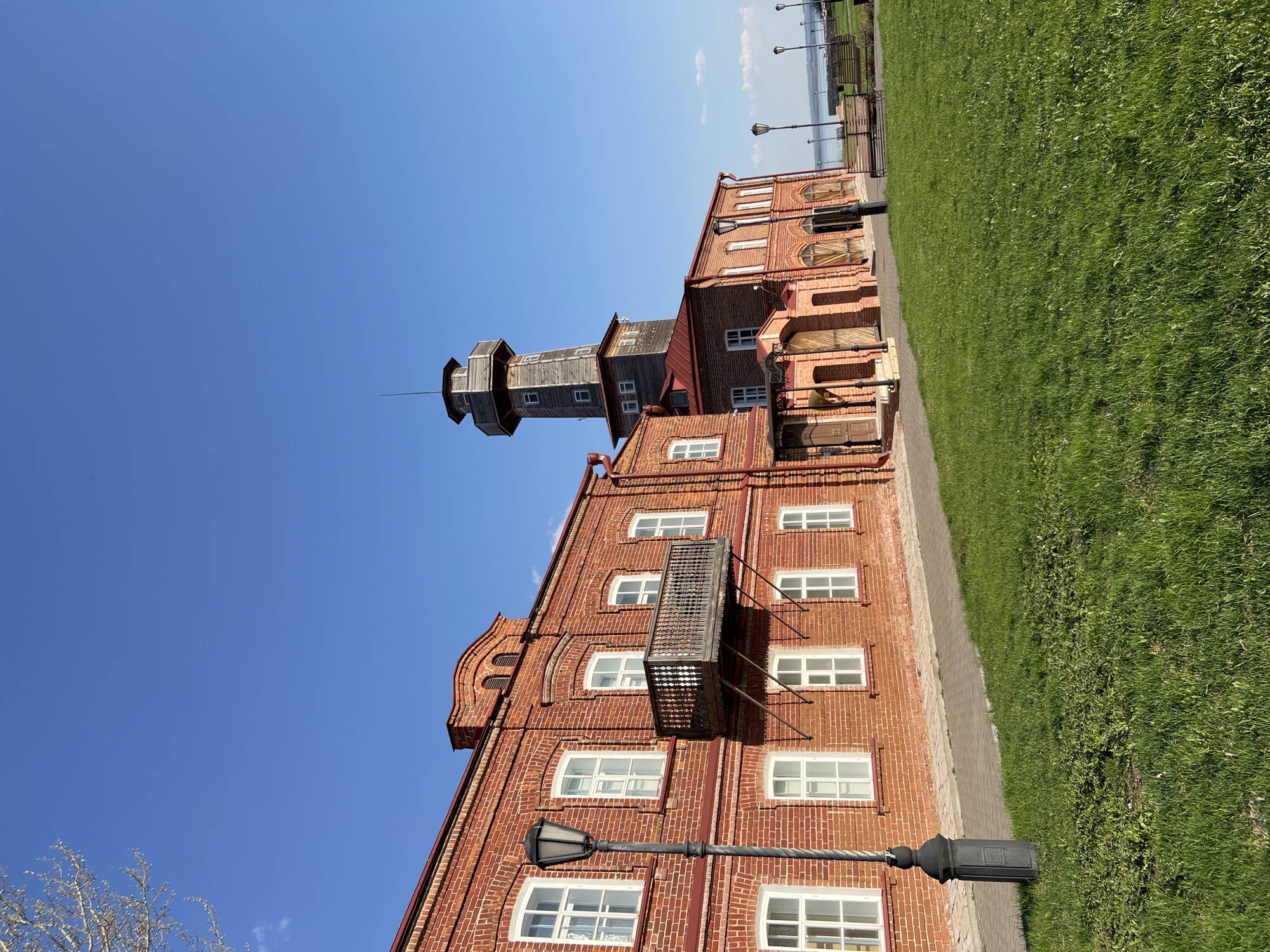 The image depicts the historic fire station in Sviyazhsk, Russia. The scene features a red brick building with multiple windows, a balcony, and a prominent wooden fire lookout tower. A pathway runs alongside the building, separating it from a grassy area. The sky is clear and blue. A lamppost stands in front of the building. Benches and street lamps can be seen in the distance. The photograph is taken at ground level with a slightly upward angle.