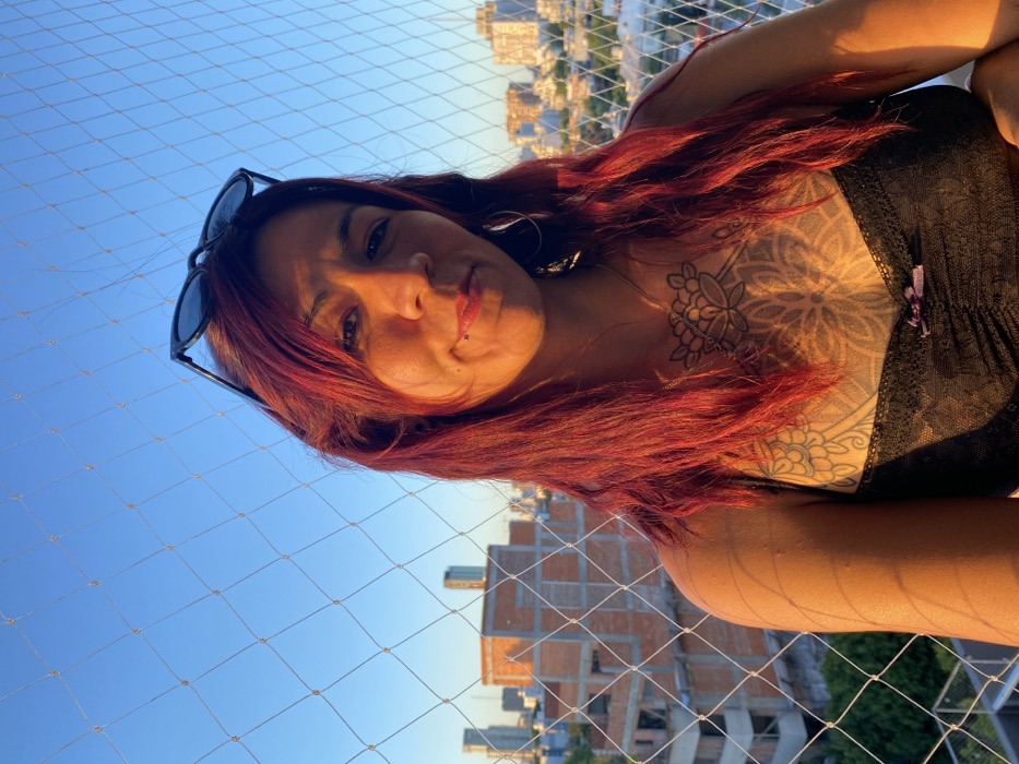 A woman with red hair and sunglasses perched on her head looks at the camera. She has a lip piercing and wears a black top that reveals a chest tattoo. The background includes a city skyline and a safety net. The lighting is warm, suggesting either sunrise or sunset.