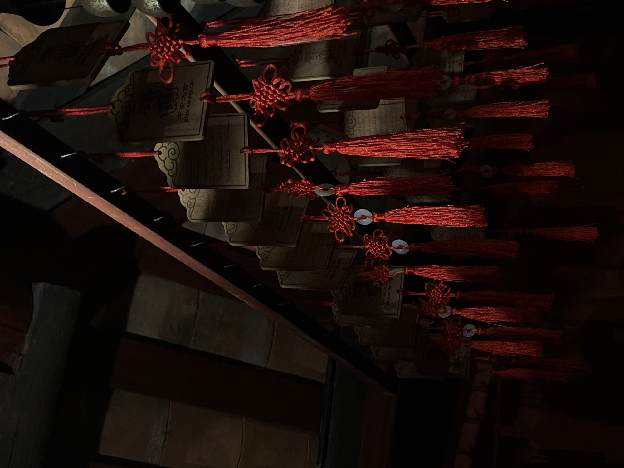 The image depicts a close-up of an interior space, likely a temple or shrine, in Hoi An, Vietnam. The scene is dominated by numerous red tassels and wooden plaques hanging from a dark wooden beam. The plaques, some of which have inscriptions in Vietnamese, are arranged in rows and appear to be prayer charms or good luck tokens. The red tassels are intricately knotted and some are adorned with small, light-colored beads. The lighting is dim, with strong shadows, suggesting the photograph was taken in low-light conditions, possibly during evening or in a shaded area. The architecture visible in the background, with its tiled roof and dark wooden beams, is characteristic of traditional Vietnamese buildings. The overall impression is one of cultural and spiritual significance, with the red color symbolizing good fortune and protection.
