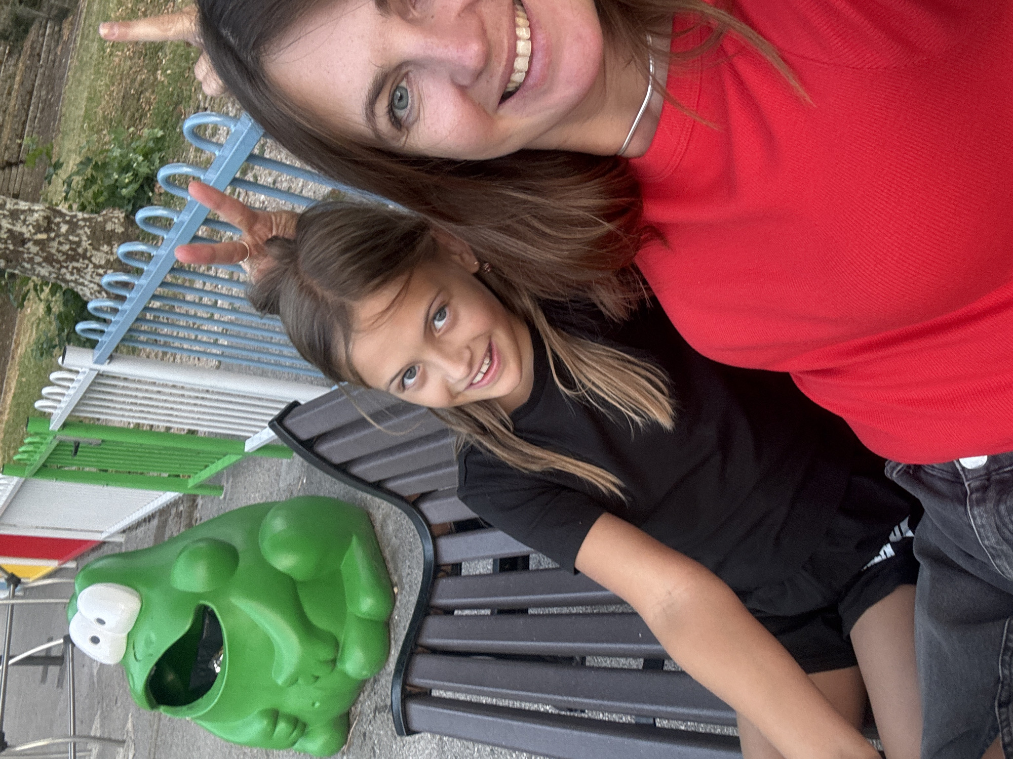This image depicts two females, likely mother and daughter, sitting on a park bench. The younger female, with blonde hair styled in a bun, is wearing a black t-shirt and black shorts. She is positioned to the left of the frame, looking towards the camera with a slight smile. To her right, an adult female with long brown hair and blue eyes, wearing a red top and a silver necklace, is also looking at the camera and smiling. Her right hand is raised, and she is making a peace sign gesture with her fingers. Behind them, a bright green frog-shaped trash can is visible on the pavement. The background consists of a metal fence, grass, and trees, suggesting an outdoor park or recreational area. The image is taken from a slightly low angle, focusing on the two individuals.