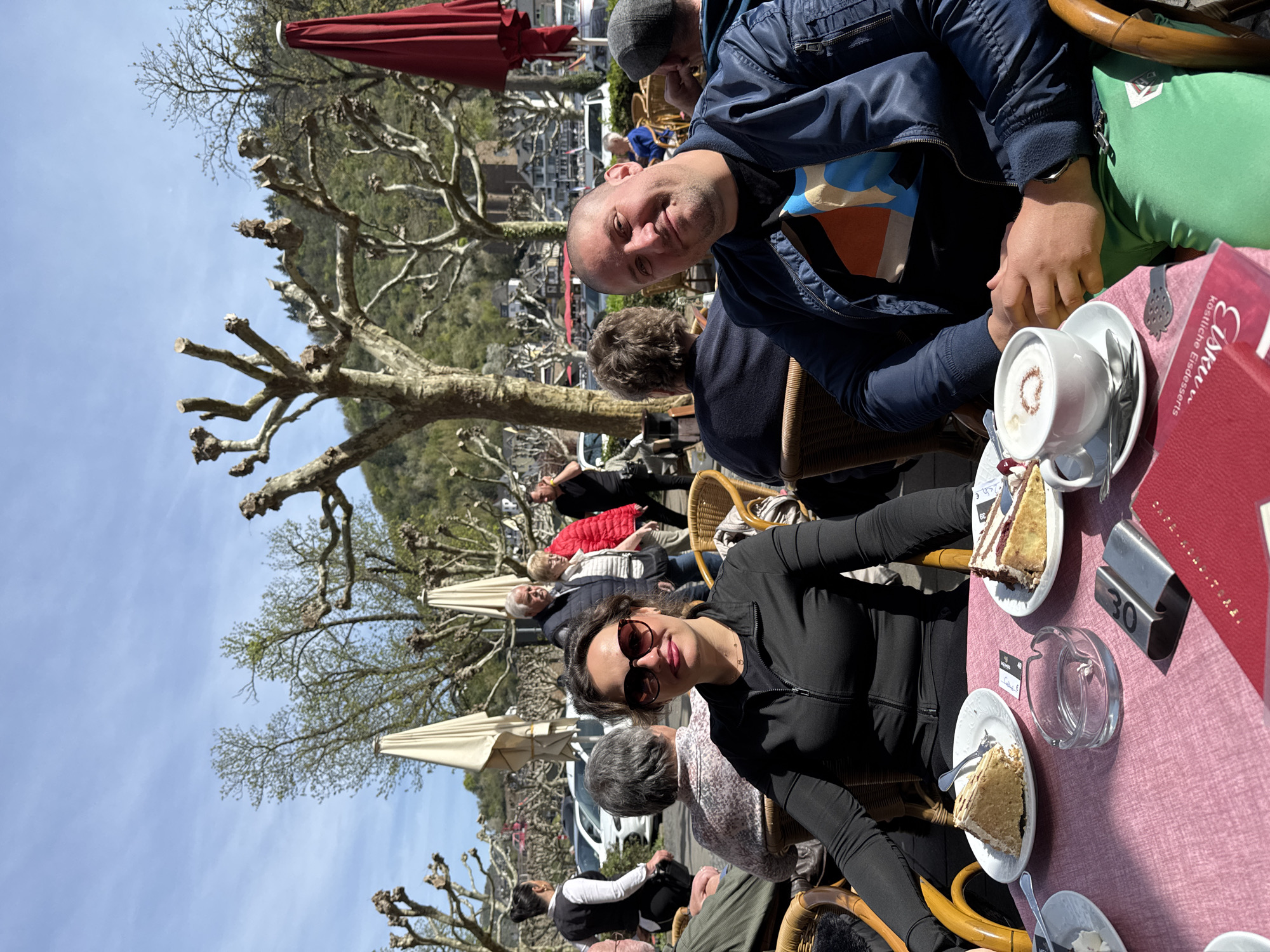 The image captures a scene at an outdoor cafe in Cochem, Germany. Two individuals are the main subjects, seated at a table covered with a pink tablecloth. A man wearing a dark blue jacket with colorful shirt underneath, sits with a light skinned male in a grey cap, while a woman in a black jacket and sunglasses sits across from them. On the table, there are plates of cake, cups with coffee, an ashtray and a red Eiskuvy Desserts menu. Behind them, there are other patrons at surrounding tables, as well as trimmed trees with bare branches, typical of a European town square. In the background, the town of Cochem and a tree-covered hillside can be seen under a clear blue sky.