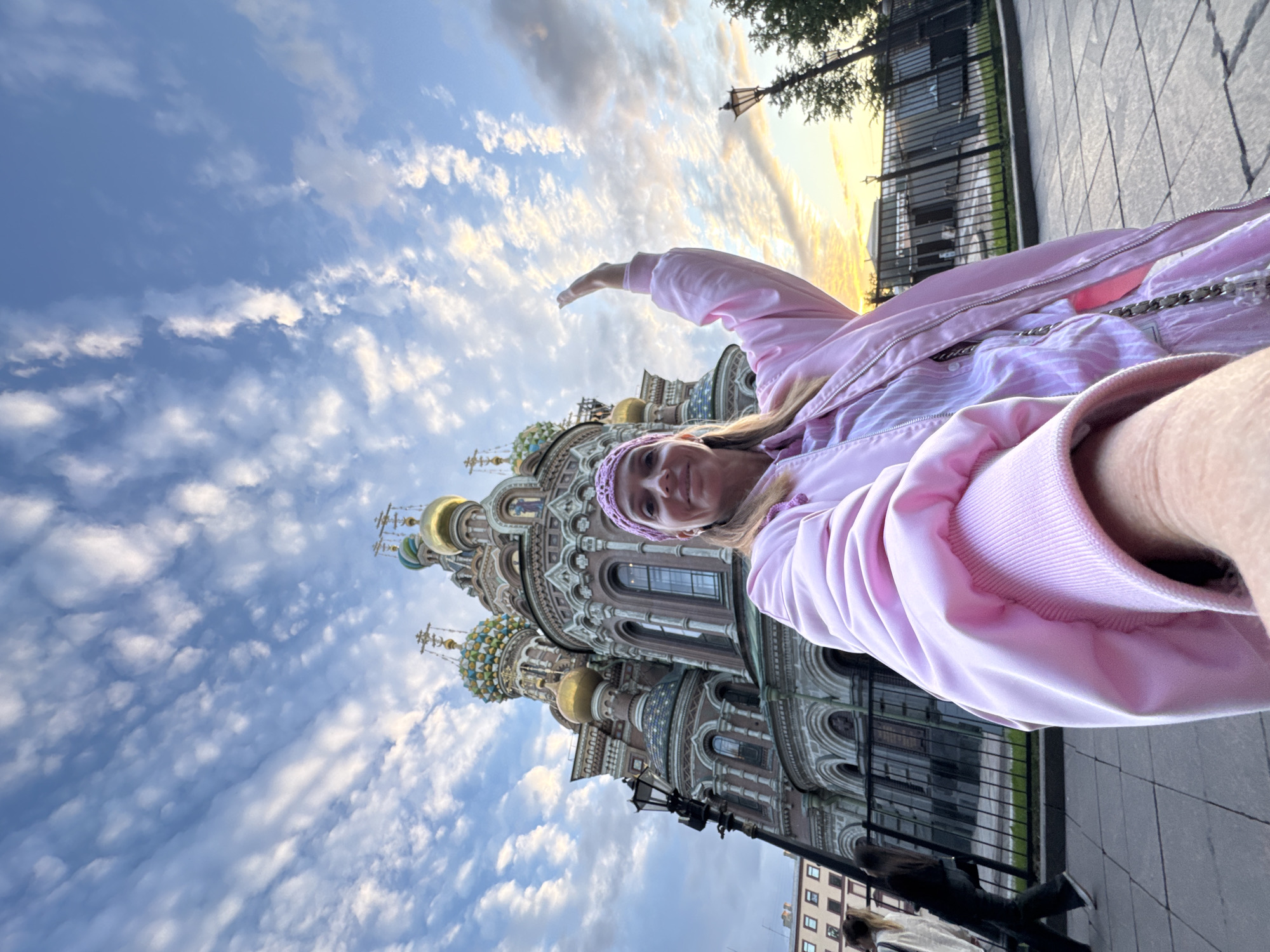 The image shows a woman taking a selfie in front of the Church of the Savior on Spilled Blood in Saint Petersburg, Russia. The church, a prominent landmark known for its colorful onion domes and intricate mosaics, is in the background, partially obscured by the woman and a fence. The woman is wearing a pink jacket and a pink headband, and her right arm is raised in a wave. The sky above is a dramatic mix of blue with wispy white clouds, suggesting either dawn or dusk. The foreground shows a paved walkway with a lamppost and some trees visible to the right. Two other individuals are walking in the background on the left side of the image. The overall scene depicts a tourist capturing a moment at a significant historical and architectural site.