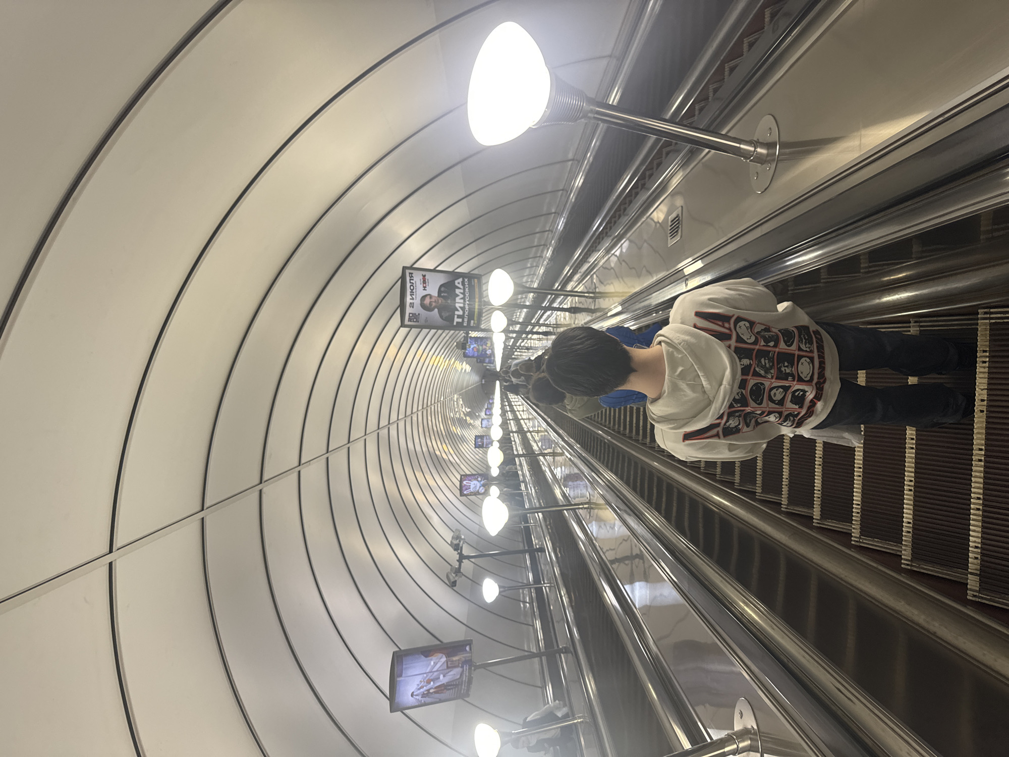 The image depicts a long, deep escalator in an underground metro station in Saint Petersburg, Russia. The scene is dominated by the immense depth of the escalator tunnel, which features a distinctive, brightly lit, arched ceiling. The ceiling is white with multiple dark, thin structural ribs curving overhead, creating a sense of rapid descent and tunnel perspective.
In the foreground, a person is seen from behind, riding down the escalator. They are wearing a white hooded sweatshirt adorned with a grid of various black and white graphic images on the back, and dark pants. Their dark hair is visible at the top of their head. Further down the escalator, other indistinct figures can be seen.
Bright, spherical light fixtures are mounted on metallic poles along the left side of the escalator, providing illumination to the tunnel. Along the same wall, several digital advertisement screens are spaced out. One prominent screen in the middle distance clearly displays Russian text: "2 ИЮЛЯ" (July 2), "ТИМА" (Tima), and "БЕЛОРУССКИХ" (Belarusian), suggesting an advertisement for an event or performer named Tima scheduled for July 2nd.
The escalator steps are dark, likely brown or black, with light-colored dividing lines, and polished metal handrails run alongside, reflecting the light. The overall impression is one of modern, functional infrastructure characteristic of deep metro systems, with a clear focus on the depth and unique architecture of the Saint Petersburg Metro. The image is oriented sideways, giving a horizontal perception to the vertical descent.